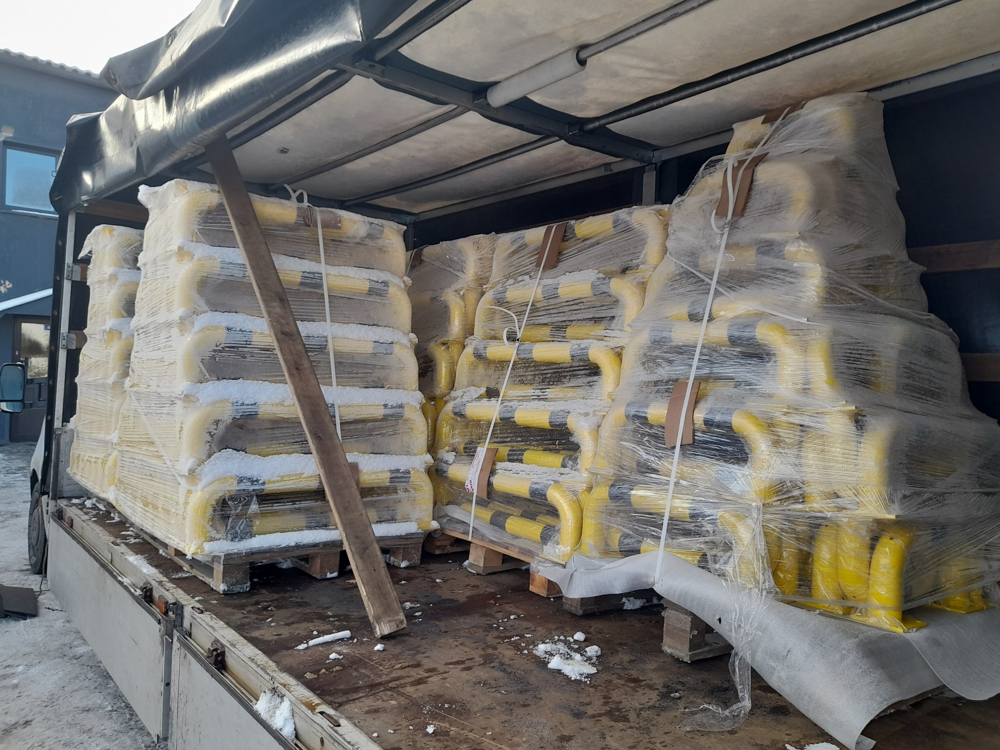 Transport słupków parkingowych w folii na paletach w przestrzeni ładunkowej samochodu dostawczego. Widoczne elementy mocujące ładunek i śnieg.