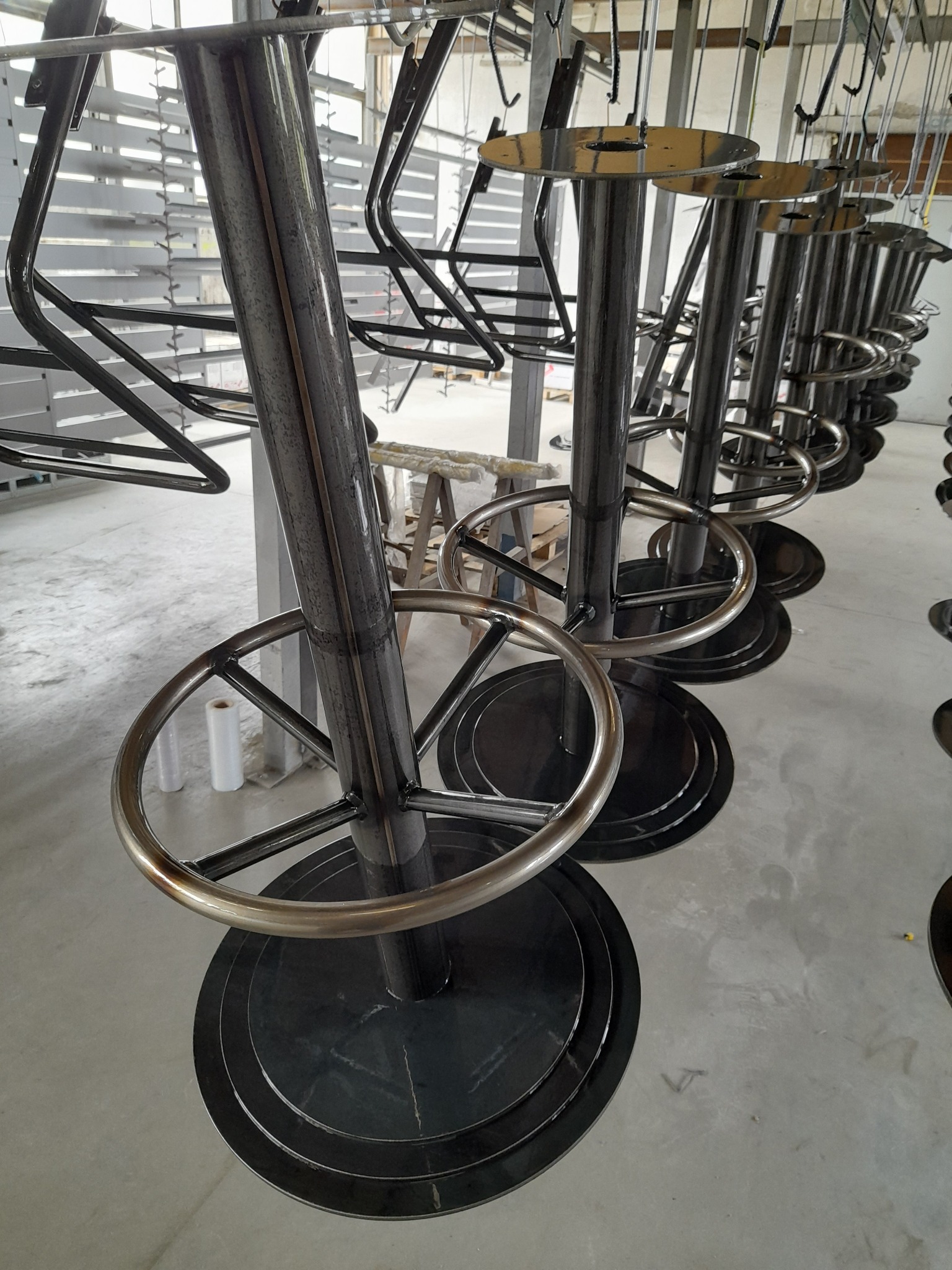 Rząd metalowych krzeseł barowych w trakcie procesu malowania proszkowego, zawieszonych na hakach w hali produkcyjnej. Widoczne elementy konstrukcyjne i surowy wygląd.