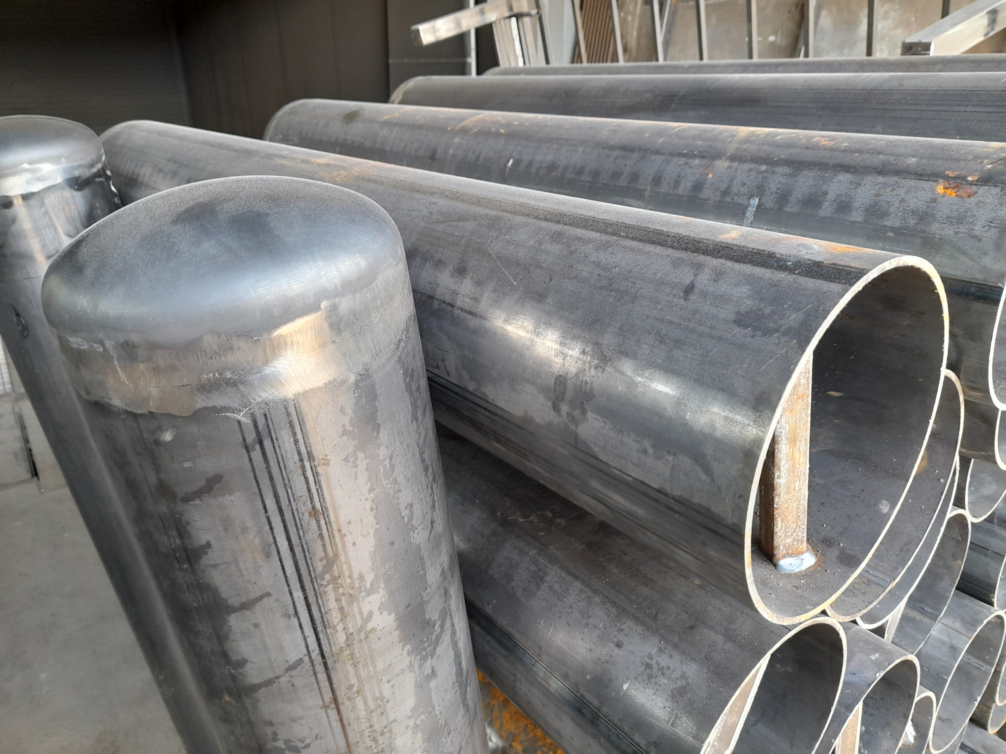 Stos rur metalowych z zaślepionymi końcami, przygotowanych do dalszej obróbki w Swarożynie. Widoczne ślady spawania i niedoskonałości powierzchni.