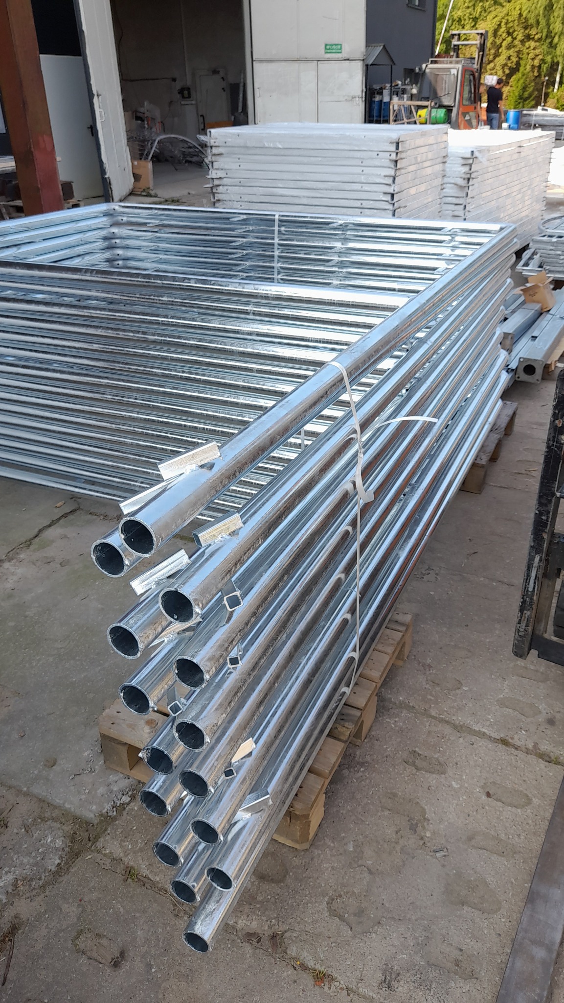 Stos ocynkowanych, metalowych słupków ogrodzeniowych na palecie, przygotowanych do transportu. W tle widoczne panele ogrodzeniowe i wózek widłowy.