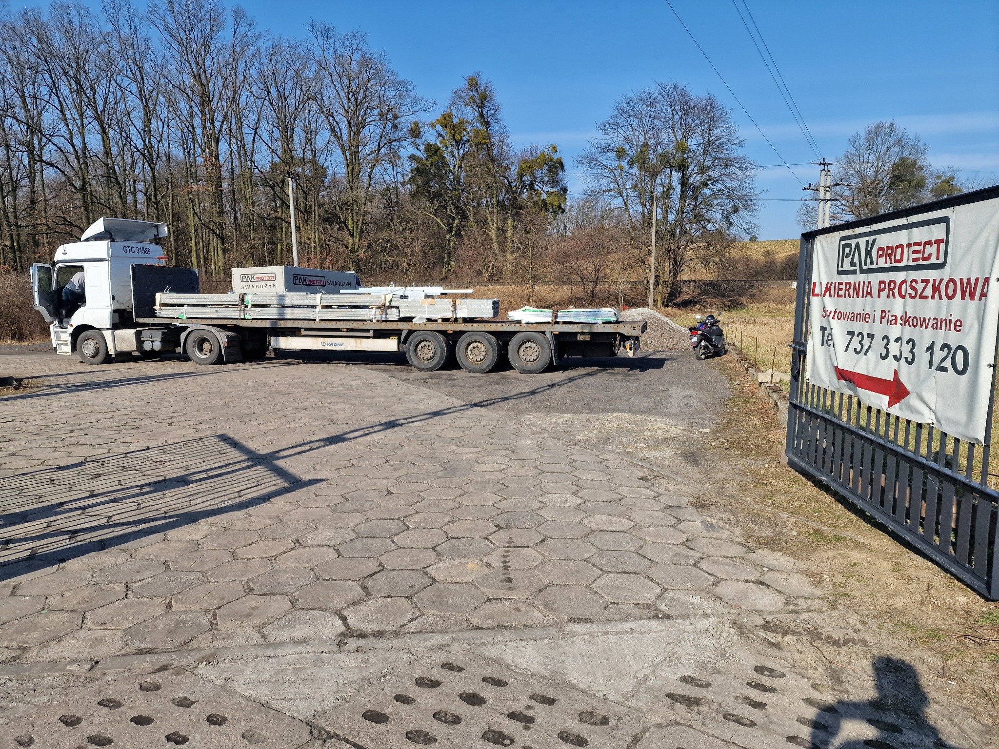 Ciężarówka z ładunkiem profili ogrodzeniowych Pak Protect Swarożyn na tle lakierni proszkowej z reklamą piaskowania. Brama wjazdowa i kostka brukowa.