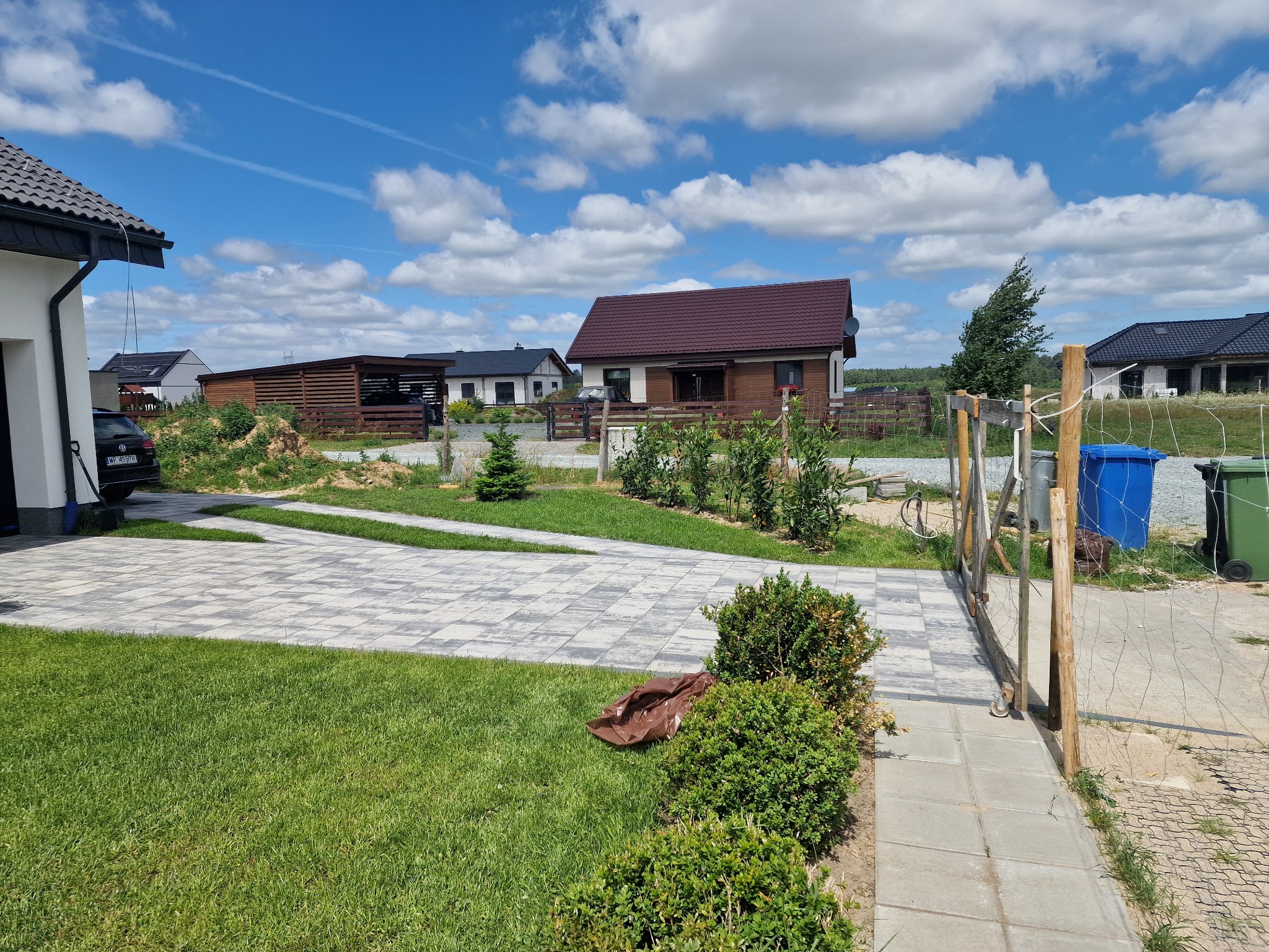 Nowoczesny dom jednorodzinny z brukowanym podjazdem i drewnianym ogrodzeniem siatkowym. Widok na zadbany trawnik i błękitne niebo z chmurami.