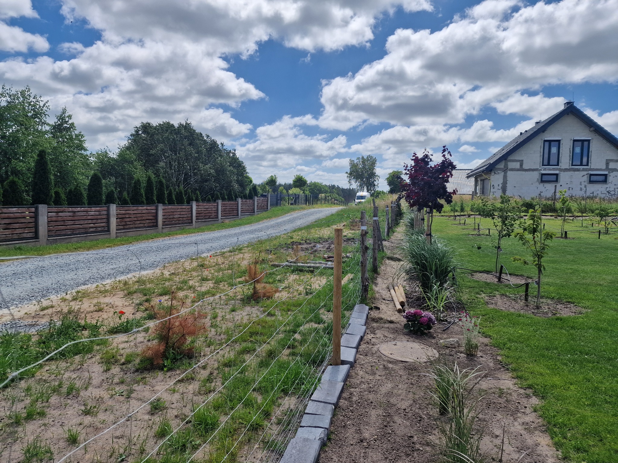 Działka z częściowo zamontowaną siatką ogrodzeniową, widoczne słupki i podmurówka z betonowych bloczków. W tle dom i droga szutrowa.