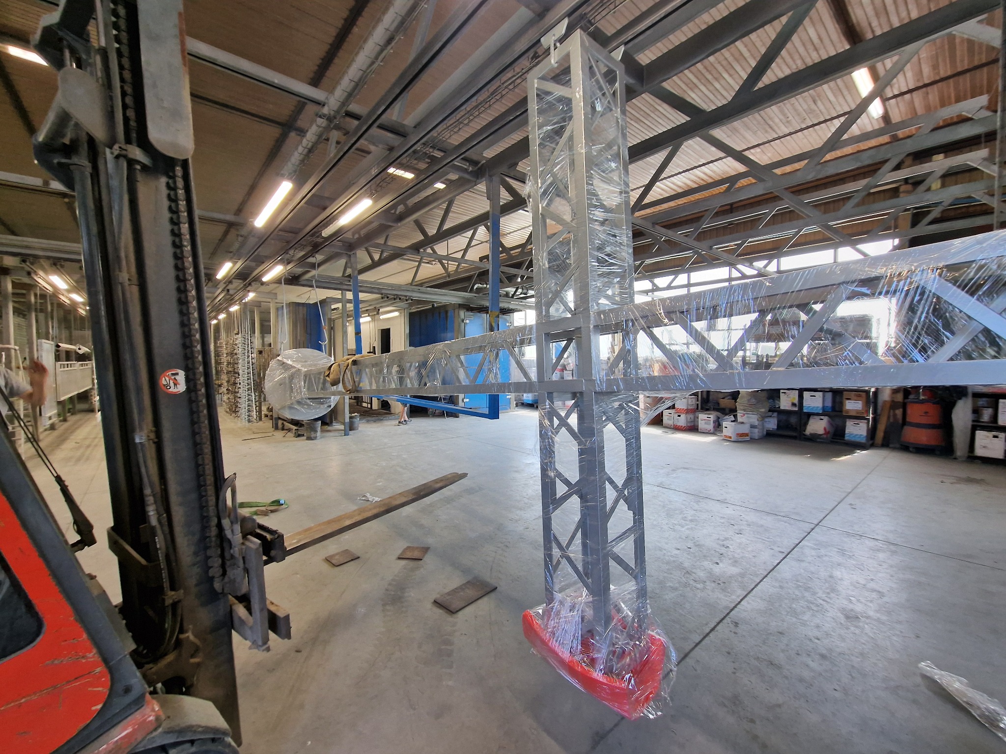 Konstrukcja metalowa, zabezpieczona folią, w hali produkcyjnej. Widoczny wózek widłowy i elementy konstrukcji dachu. Jasne oświetlenie, betonowa podłoga.