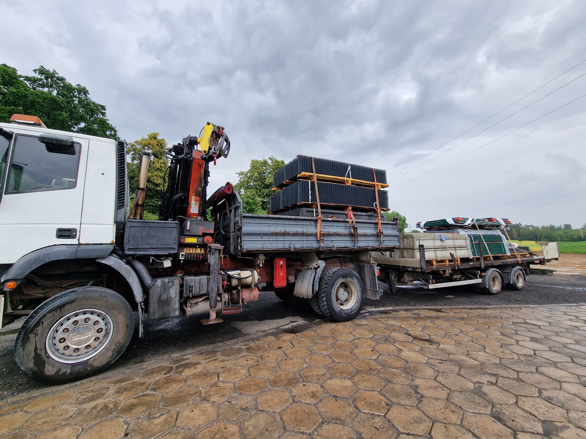 Ciężarówka z HDS przewozi panele ogrodzeniowe na paletach. Widok z boku, na pierwszym planie bruk, w tle zieleń i pochmurne niebo.