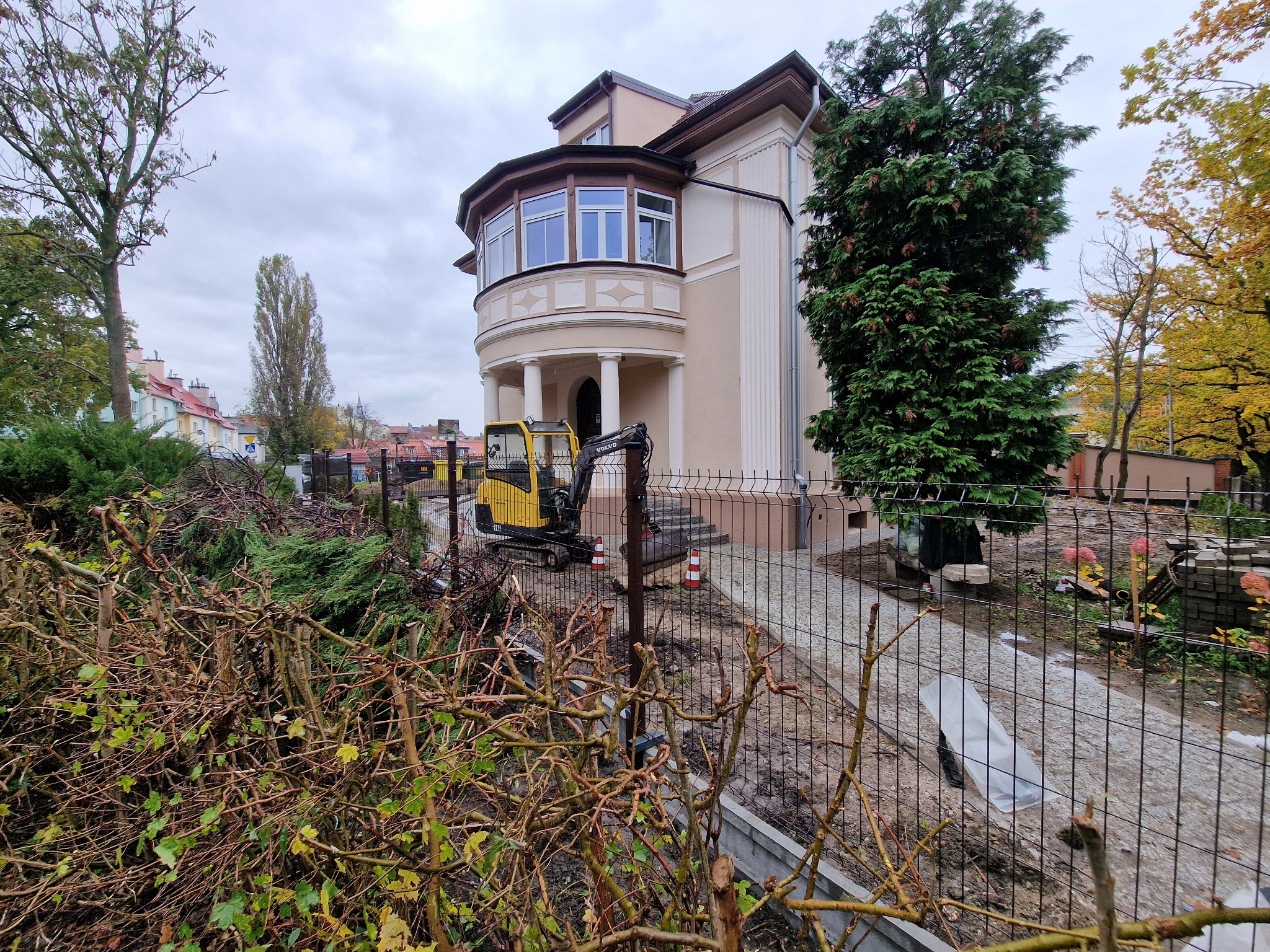 Żółta minikoparka przy nowym, ciemnym ogrodzeniu panelowym na tle beżowej willi z kolumnami i zieleni. Widok zza krzewów.