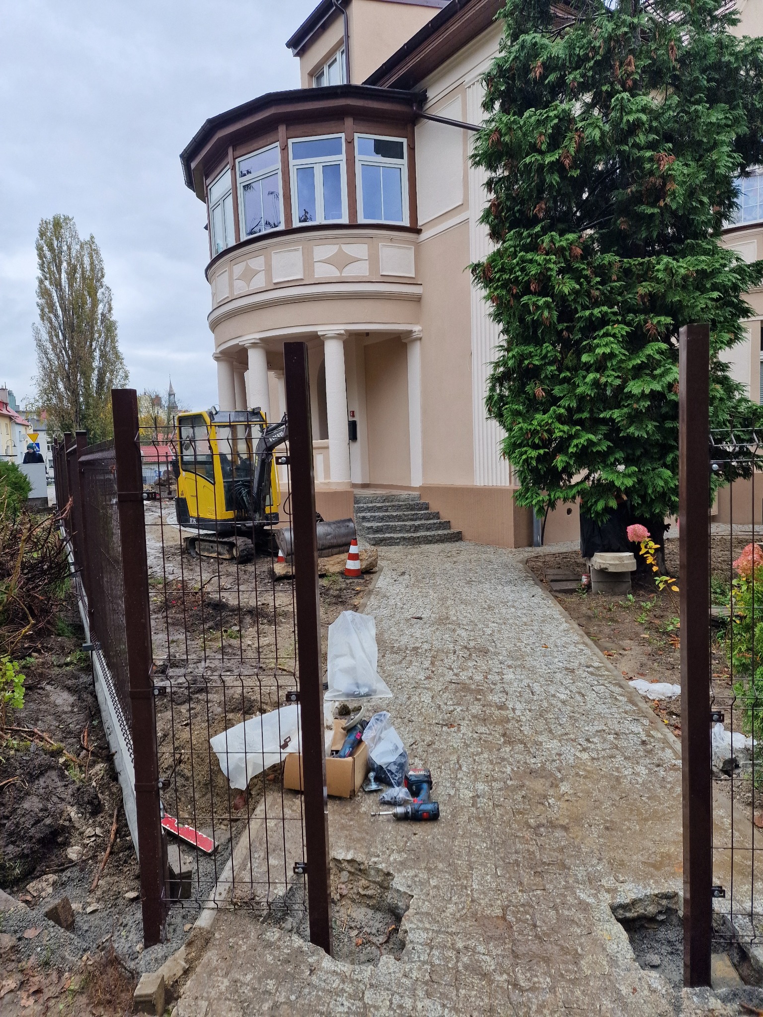 Montaż brązowego ogrodzenia panelowego na posesji z użyciem mini koparki. Widoczne narzędzia i elementy montażowe. Tło stanowi budynek z kolumnami i zielone drzewo.
