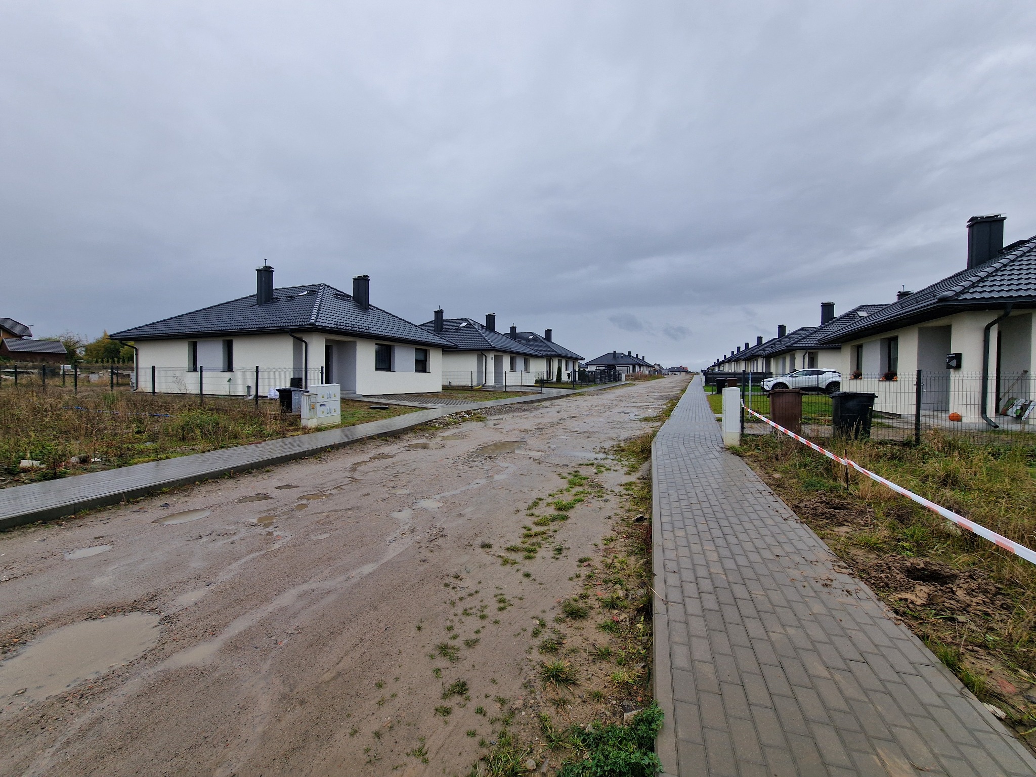 Szereg identycznych, nowych domów jednorodzinnych z czarnymi dachami, widziane z perspektywy ulicy w pochmurny dzień. Widoczne chodniki z kostki i błotnista droga.
