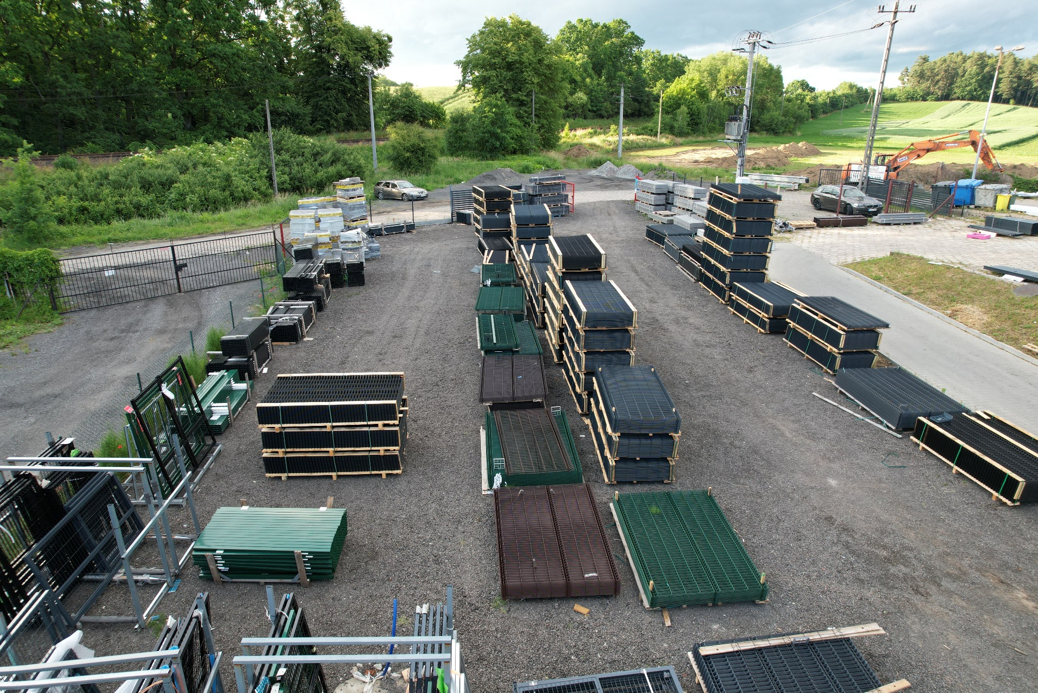 Szeroki kadr z lotu ptaka na składowisko paneli ogrodzeniowych w różnych kolorach, ułożonych w stosy na żwirowym podłożu. W tle zieleń drzew i koparka.