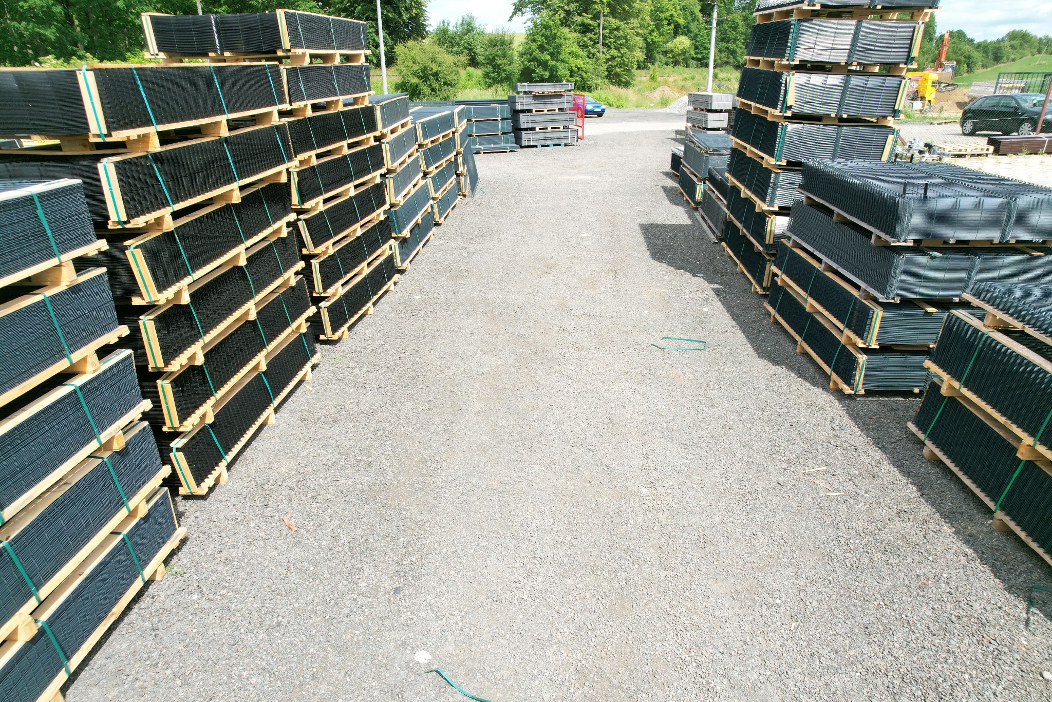 Stosy paneli ogrodzeniowych na paletach, ułożone w rzędach na żwirowym placu. Widok z góry, w tle zieleń drzew i zaparkowane pojazdy.