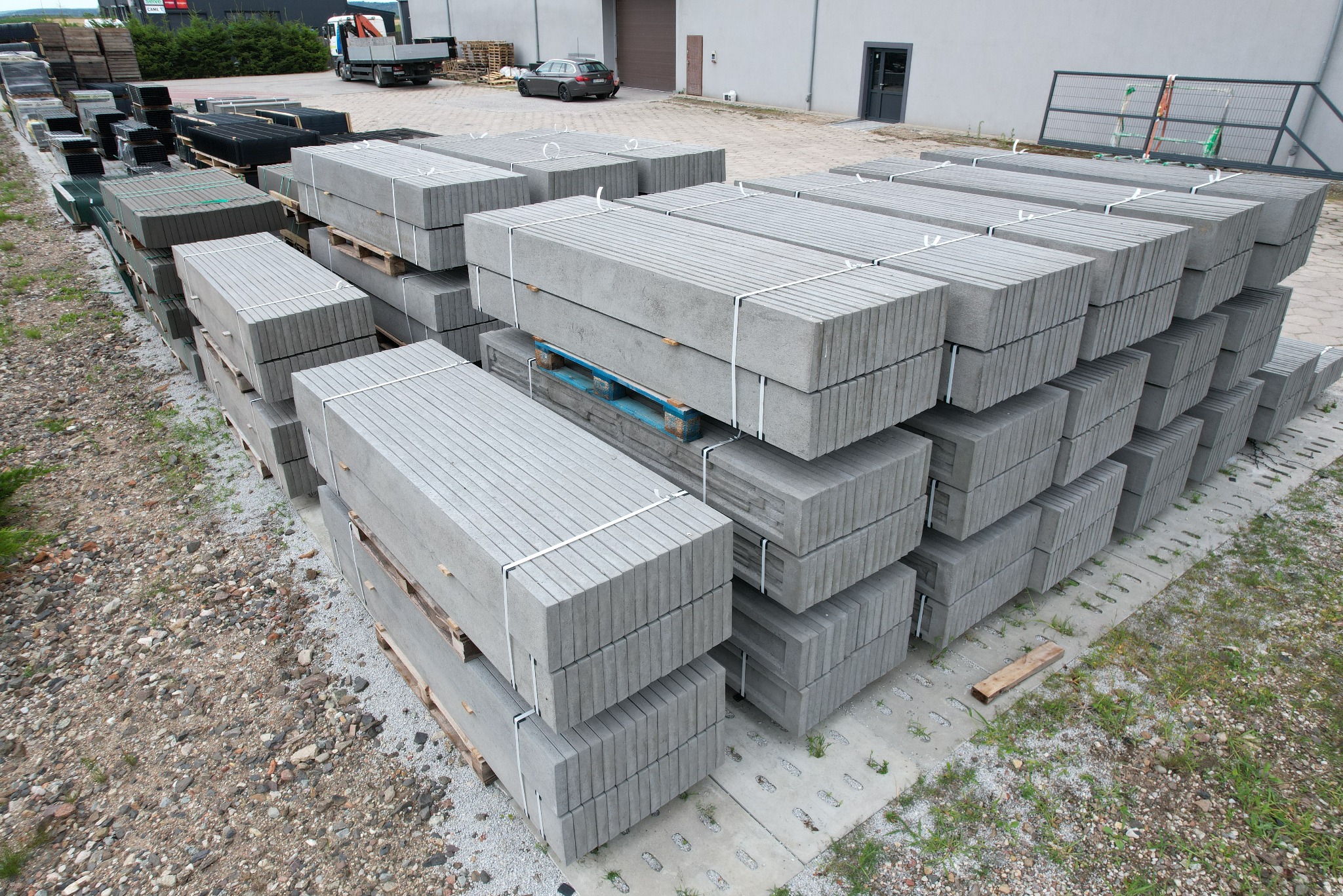 Szare, betonowe obrzeża palisadowe ułożone w stosy na paletach, przygotowane do transportu. Widoczne tło z budynkiem i pojazdami.