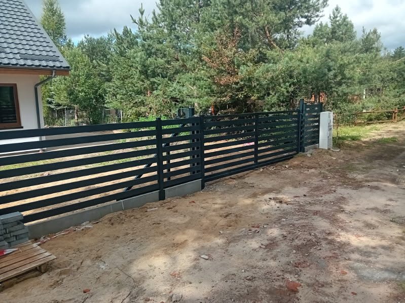 Nowoczesne, poziome, czarne ogrodzenie metalowe z bramą przesuwną na tle domu i drzew. Widoczne betonowe fundamenty i elementy montażowe. Ujęcie z poziomu gruntu.