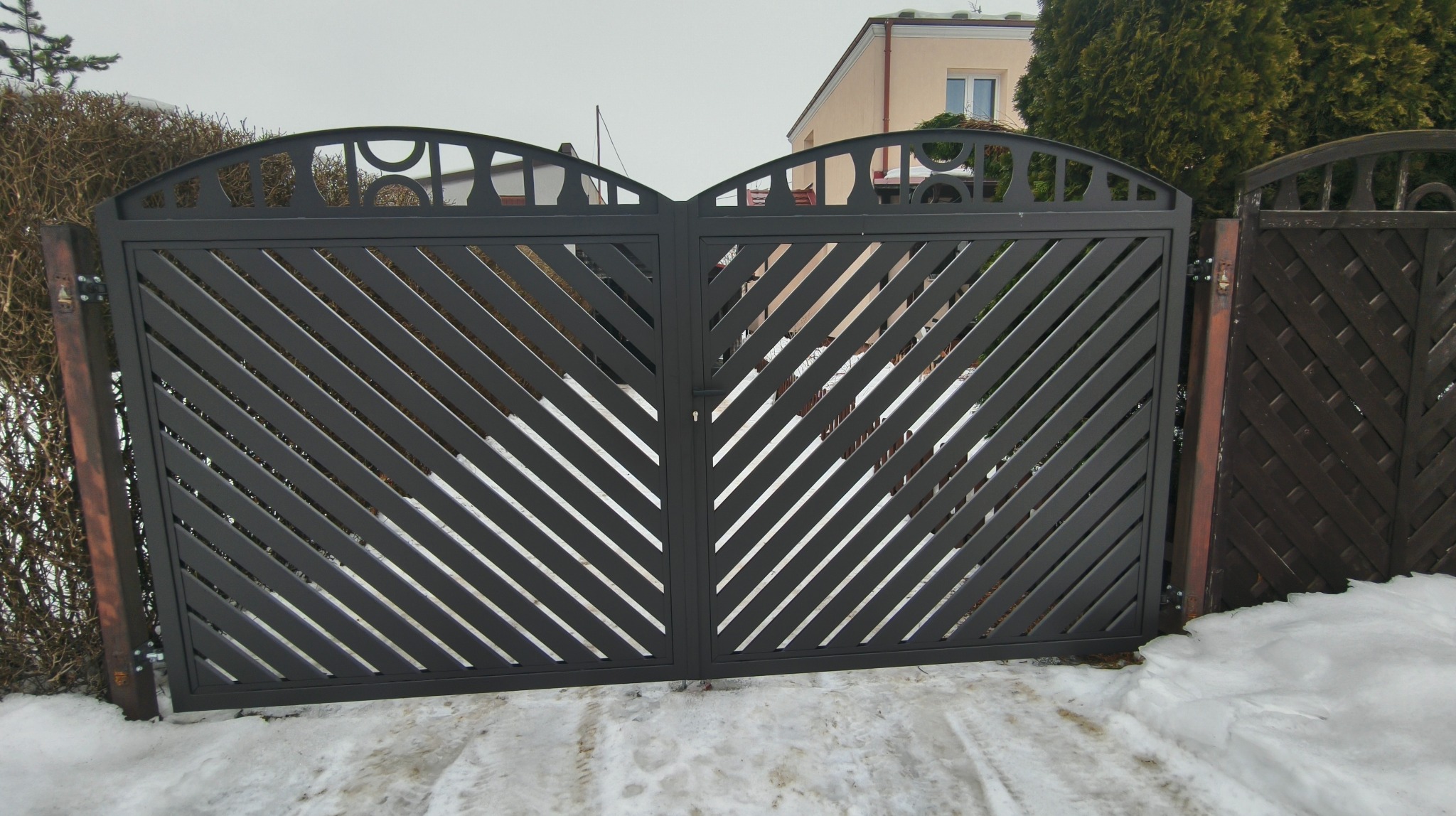 Nowoczesna, grafitowa brama wjazdowa z ukośnymi szczeblami i dekoracyjnym łukiem, zamontowana na tle zimowego krajobrazu z budynkiem w oddali.