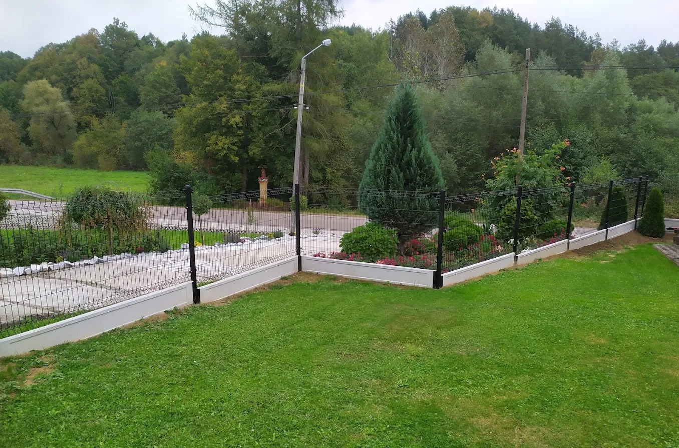 Nowoczesne ogrodzenie panelowe z podmurówką, otoczone zielenią. Widok na zadbany ogród i drzewa w tle. Słup oświetleniowy w oddali.