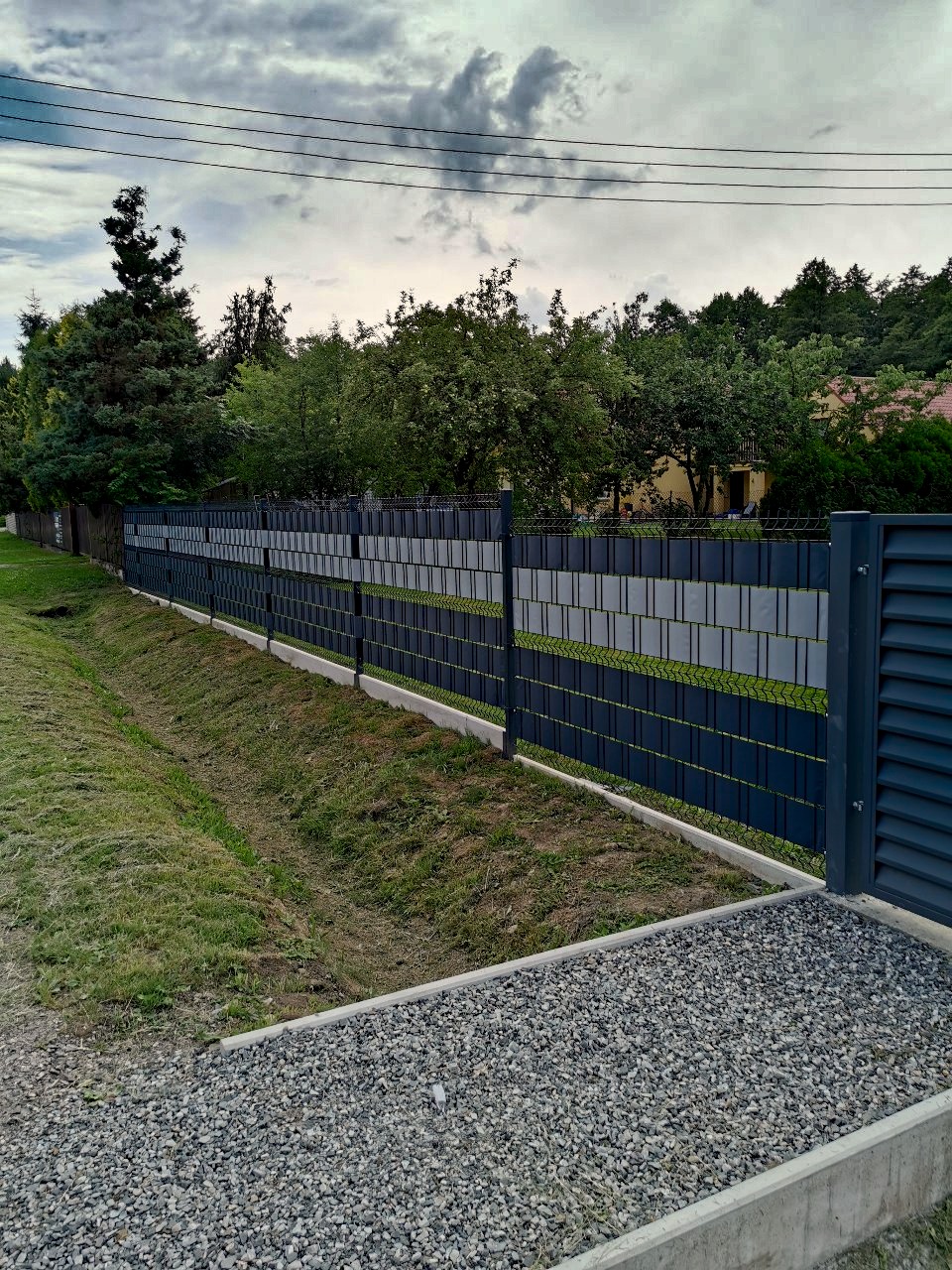 Nowoczesne ogrodzenie panelowe z bramą w kolorze grafitowym, z betonową podmurówką, na tle zieleni i pochmurnego nieba. Widok z zewnątrz posesji.