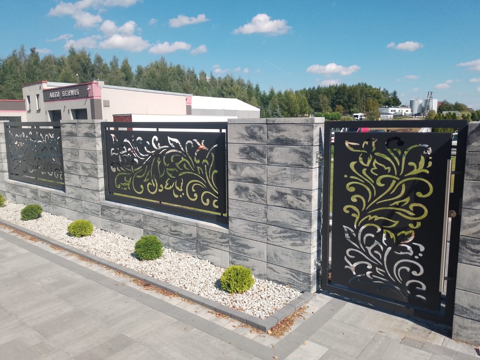 Nowoczesne ogrodzenie z paneli z wycinanym wzorem roślinnym, osadzone na murku z szarej kostki. Małe krzewy i biały żwir u podstawy. W tle budynek Auto Serwisu.