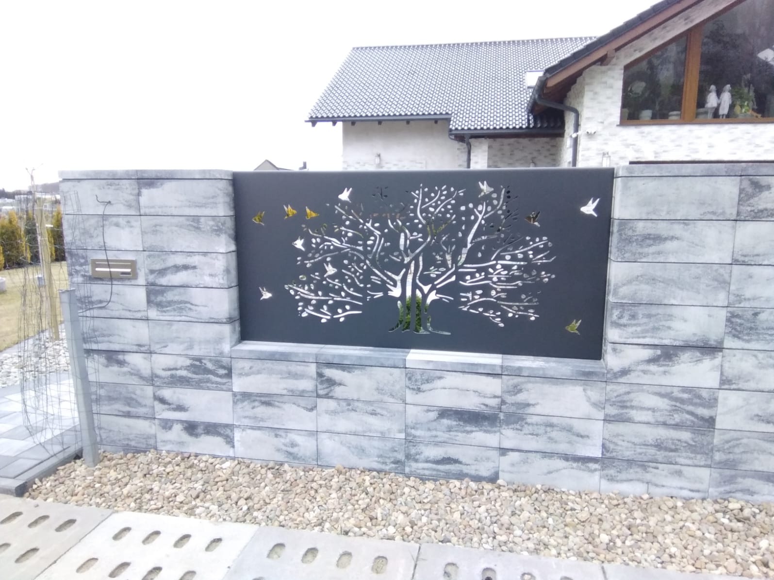 Dekoracyjny panel ogrodzeniowy z motywem drzewa i ptaków, wkomponowany w mur z szarej kostki. Nowoczesny design z elementami ażurowymi.