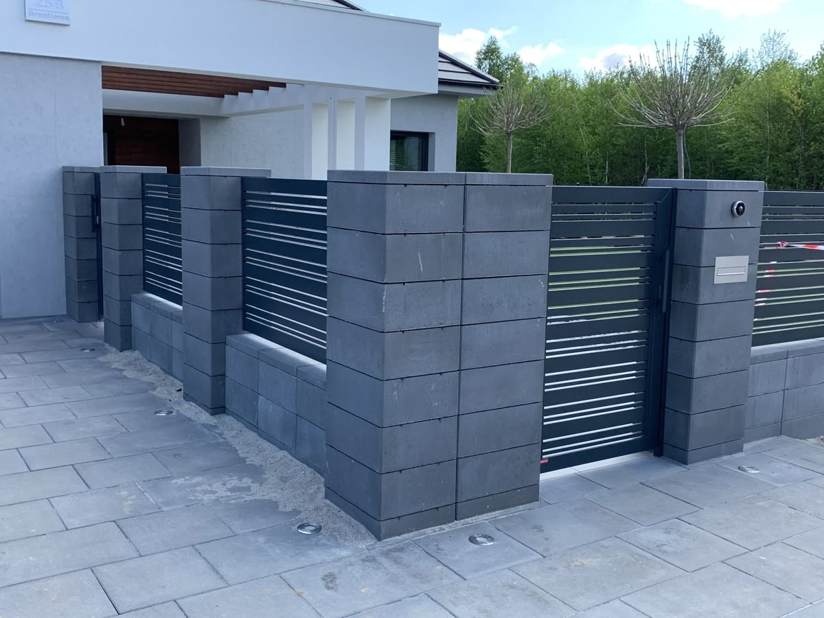 Nowoczesna brama wjazdowa z poziomymi, białymi listwami, osadzona w szarych, betonowych słupkach. Widoczny fragment domu i kostka brukowa.