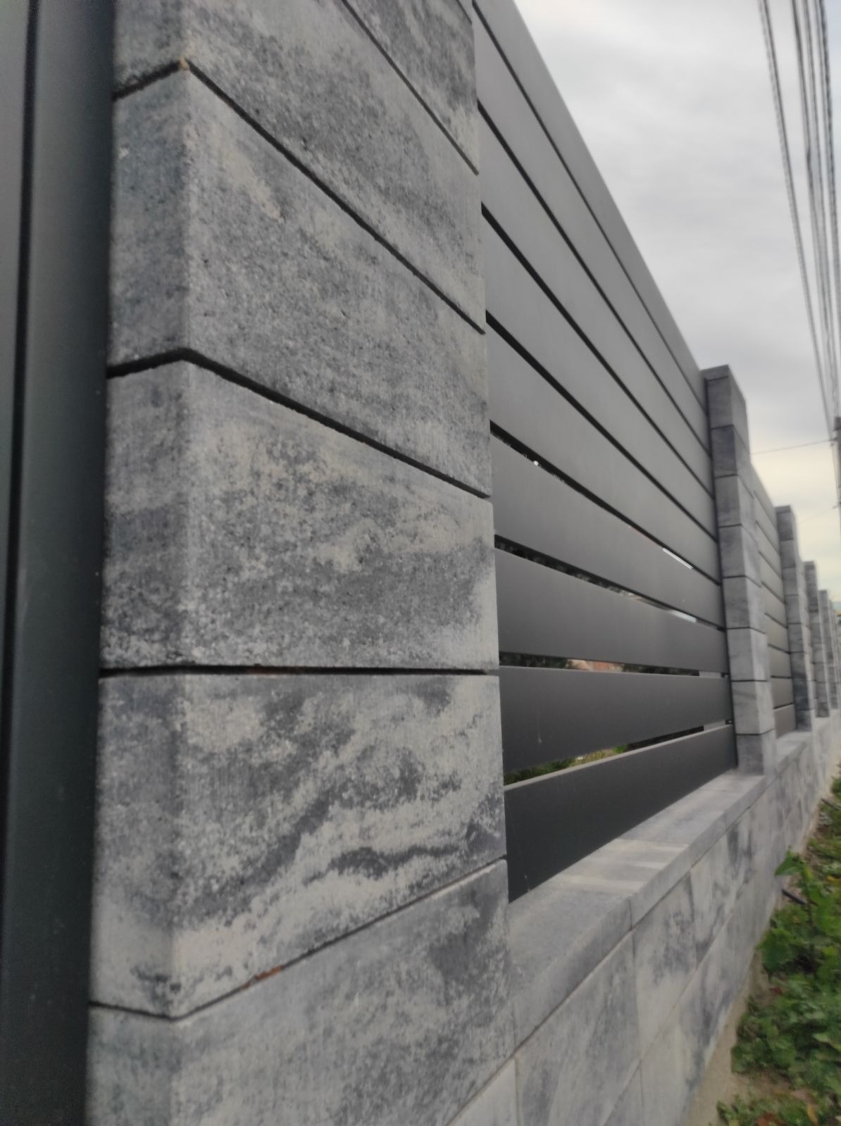 Nowoczesne ogrodzenie z szarego betonu architektonicznego i metalowych paneli w Swarożynie. Widok z bliska na słupki i przęsła, z perspektywą wzdłuż linii ogrodzenia.