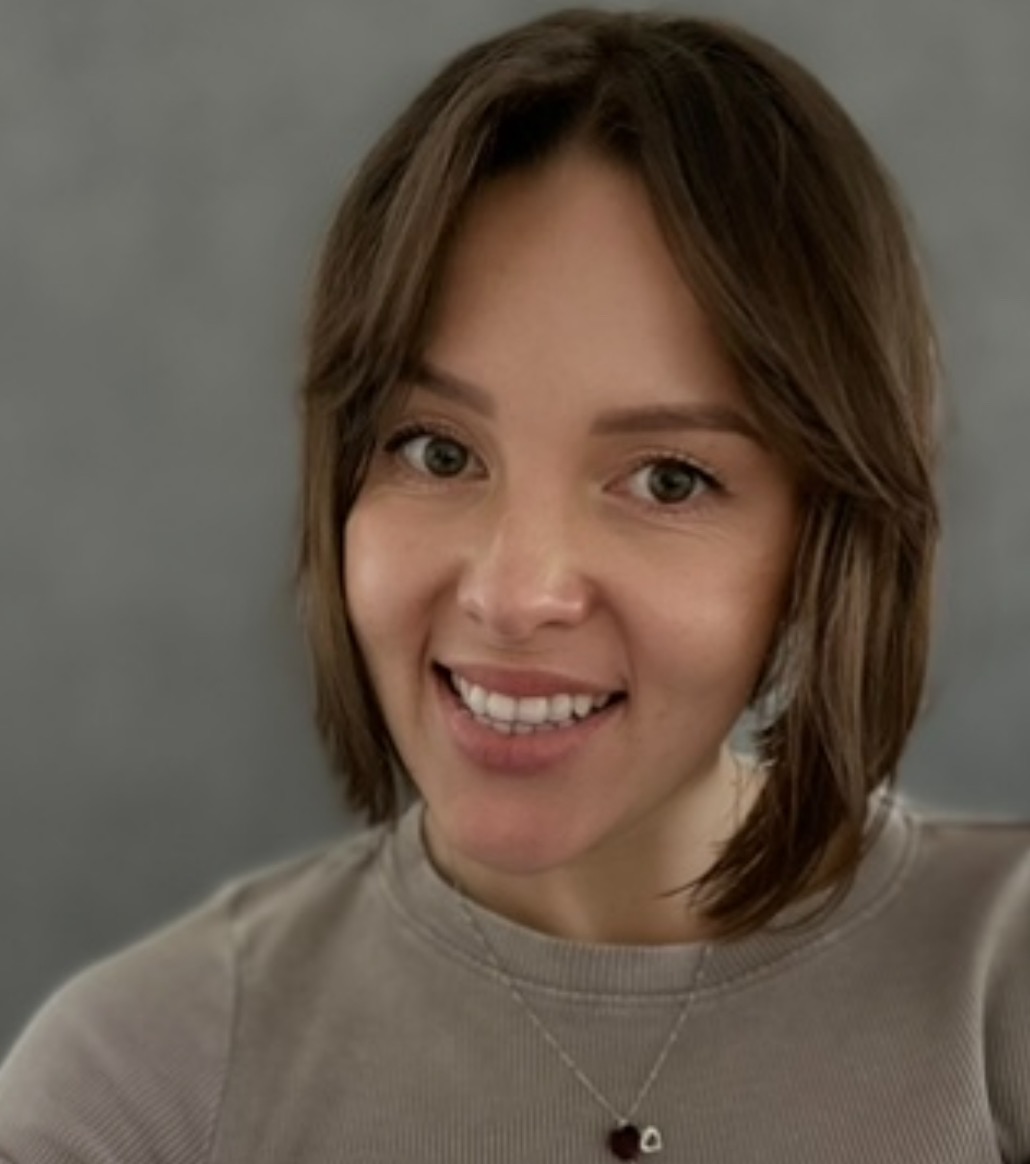 Uśmiechnięta kobieta z brązowymi włosami do ramion, w szarej bluzce i srebrnym naszyjniku z zawieszką w kształcie serca, na rozmytym szarym tle.