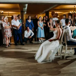 Para Młoda podczas oczepin siedzi na krzesłach, odpowiadając na pytania gości weselnych. W tle zgromadzeni goście z tabliczkami 'Ja' i 'Ona'.