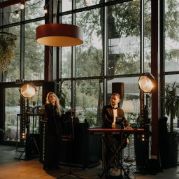 RECORD CONSULTING GROUP SPÓŁKA Z OGRANICZONĄ ODPOWIEDZIALNOŚCIĄ - Duet muzyczny: wokalistka z mikrofonem i klawiszowiec w smokingu, oświetleni reflektorami, na tle okien w przestronnym wnętrzu.