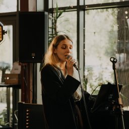 RECORD CONSULTING GROUP SPÓŁKA Z OGRANICZONĄ ODPOWIEDZIALNOŚCIĄ - Wokalistka z blond włosami śpiewa do mikrofonu na tle okna i sprzętu nagłośnieniowego. Scena oświetlona ciepłym światłem, widoczny statyw i głośnik.