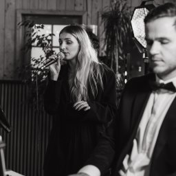 RECORD CONSULTING GROUP SPÓŁKA Z OGRANICZONĄ ODPOWIEDZIALNOŚCIĄ - Czarno-białe zdjęcie zespołu muzycznego: wokalistka z mikrofonem i pianista w smokingu, grający na pianinie. W tle drewniana ściana i oświetlenie sceniczne.