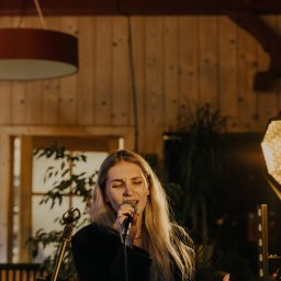 RECORD CONSULTING GROUP SPÓŁKA Z OGRANICZONĄ ODPOWIEDZIALNOŚCIĄ - Blondwłosa wokalistka śpiewa do mikrofonu na scenie z drewnianym tłem, oświetlona ciepłym światłem reflektorów. Widoczny statyw mikrofonowy i fragment oświetlenia scenicznego.