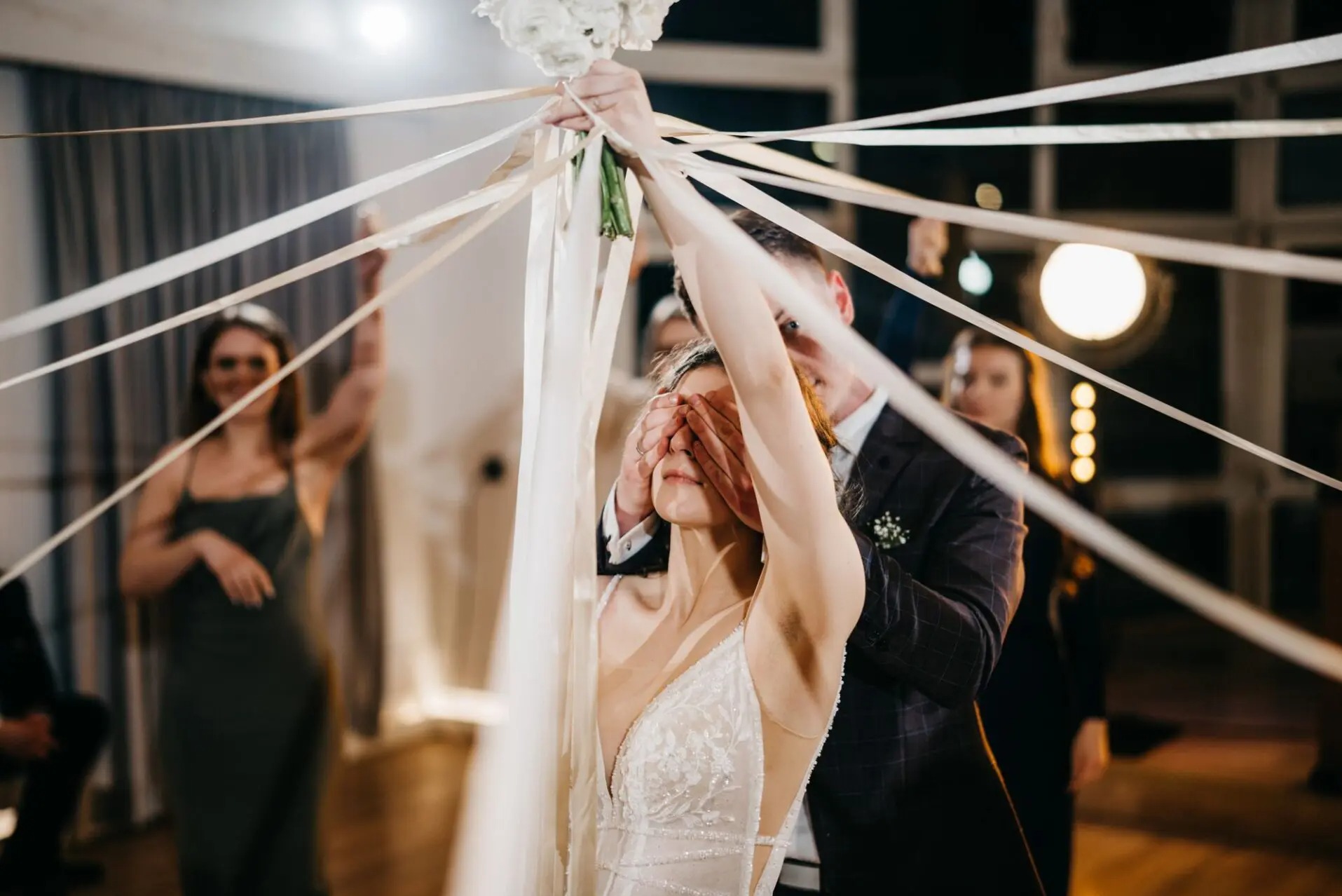 Panna młoda z zasłoniętymi oczami, trzymająca wstążki przymocowane do bukietu ślubnego, w tle goście weselni i dekoracje świetlne.