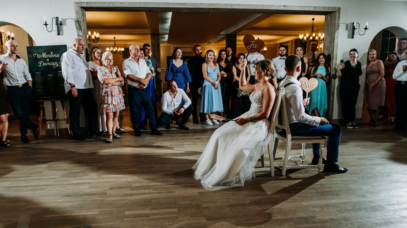 Para Młoda podczas oczepin siedzi na krzesłach, odpowiadając na pytania gości weselnych. W tle zgromadzeni goście z tabliczkami 'Ja' i 'Ona'.