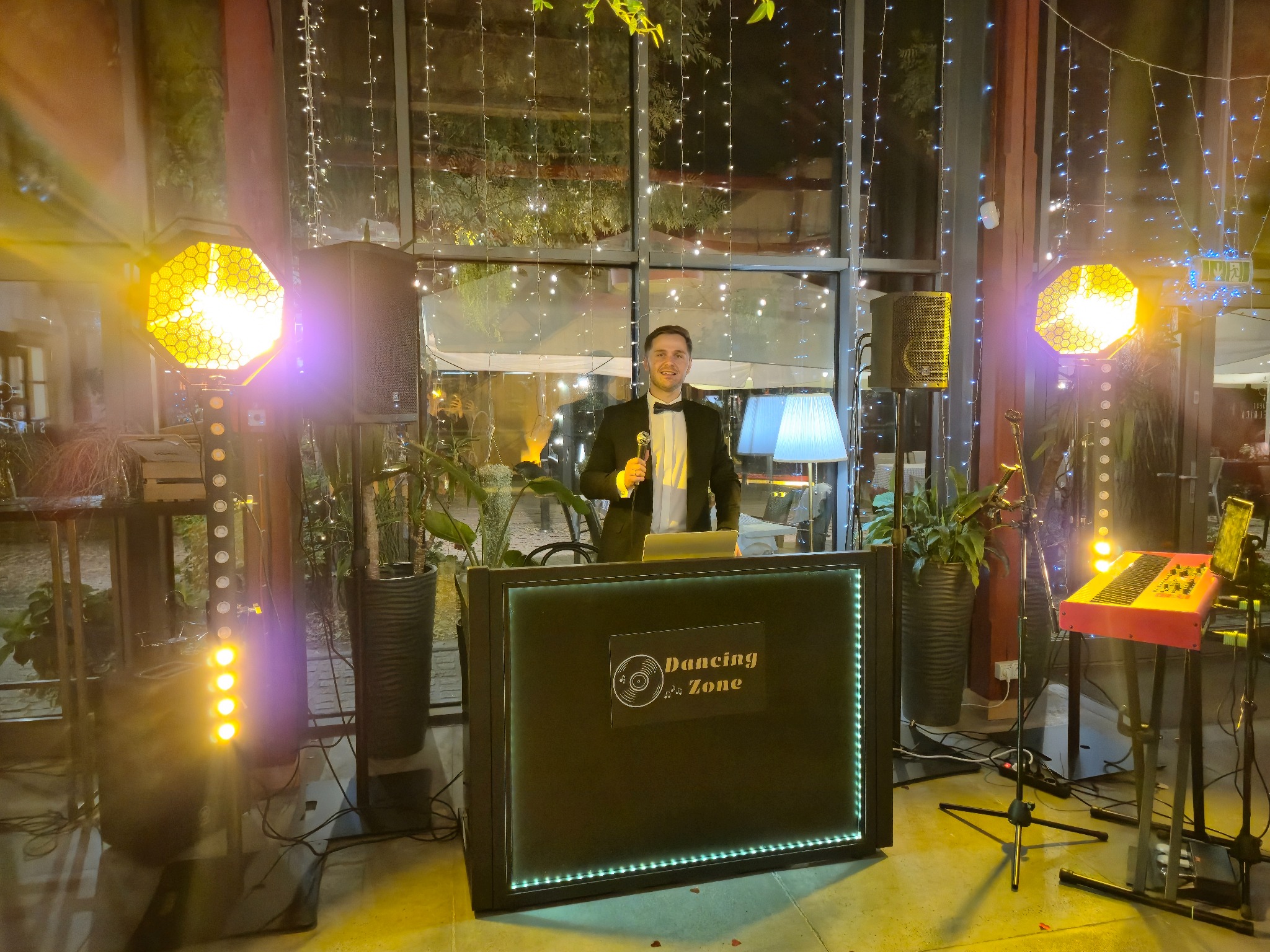 DJ w smokingu za podświetlonym stanowiskiem 'Dancing Zone', mikrofon w dłoni, w tle dekoracje świetlne i rośliny doniczkowe, widoczne elementy nagłośnienia i keyboard.