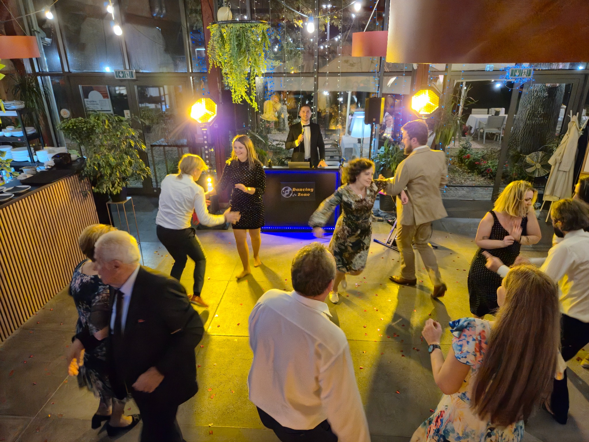 DJ w czarnym smokingu za podświetlonym stanowiskiem 'Dancing in Zone' gra dla tańczących gości w sali z dekoracjami świetlnymi i roślinnymi.
