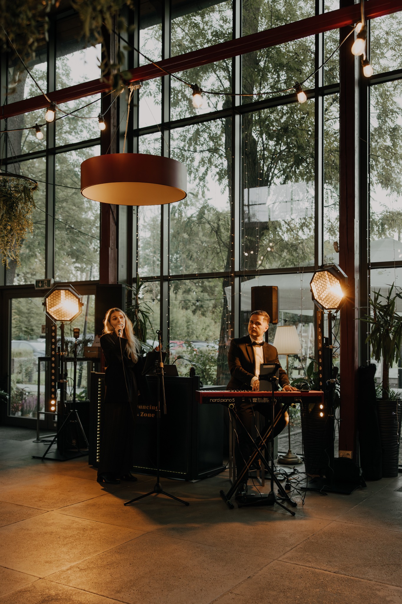Duet muzyczny: wokalistka z mikrofonem i klawiszowiec w smokingu, oświetleni reflektorami, na tle okien w przestronnym wnętrzu.