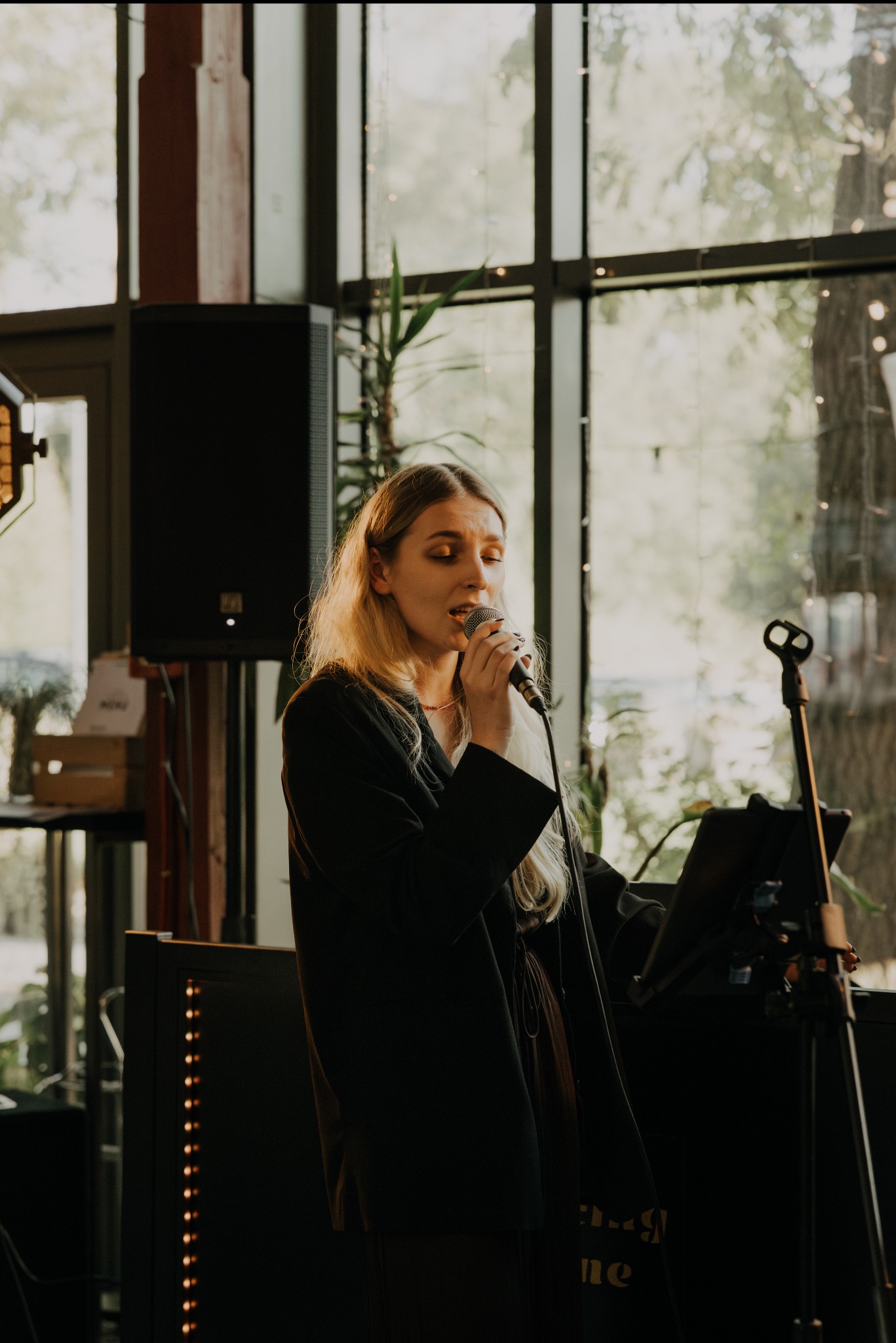 Wokalistka z blond włosami śpiewa do mikrofonu na tle okna i sprzętu nagłośnieniowego. Scena oświetlona ciepłym światłem, widoczny statyw i głośnik.
