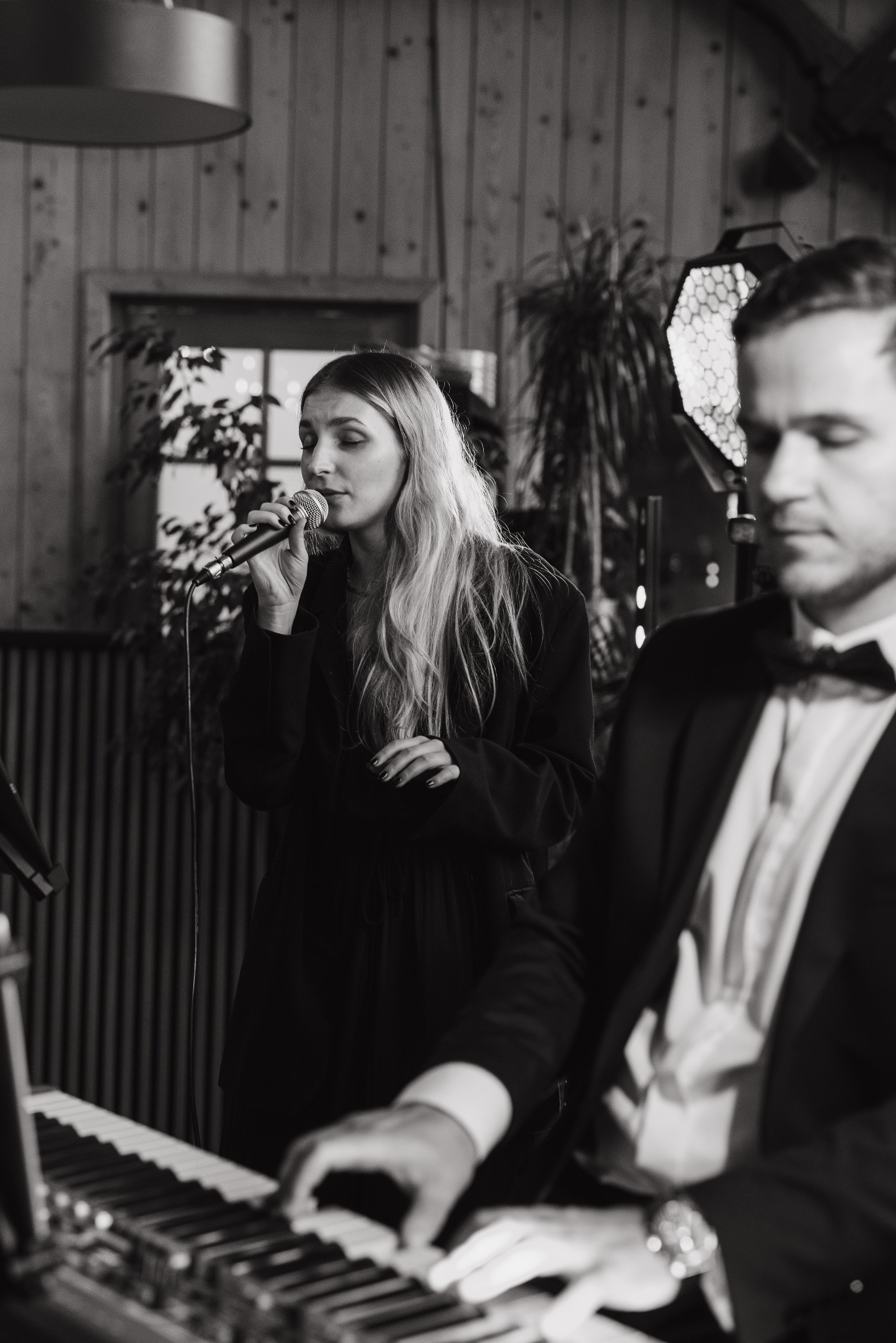 Czarno-białe zdjęcie zespołu muzycznego: wokalistka z mikrofonem i pianista w smokingu, grający na pianinie. W tle drewniana ściana i oświetlenie sceniczne.