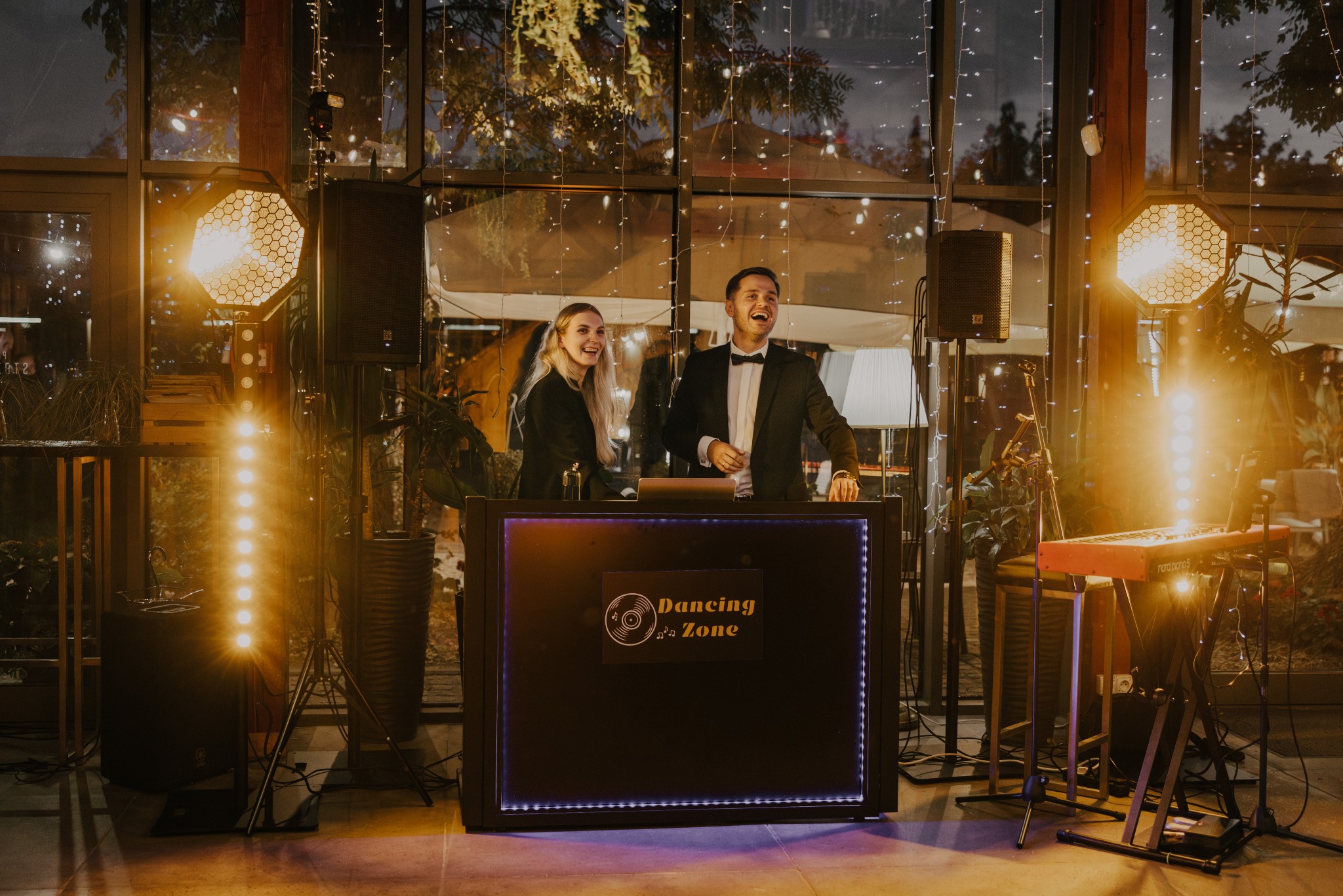 Uśmiechnięty DJ i wokalistka za konsoletą 'Dancing Zone', oświetlenie sceniczne, keyboard. Dekoracja świetlna w tle, sala z dużymi oknami.