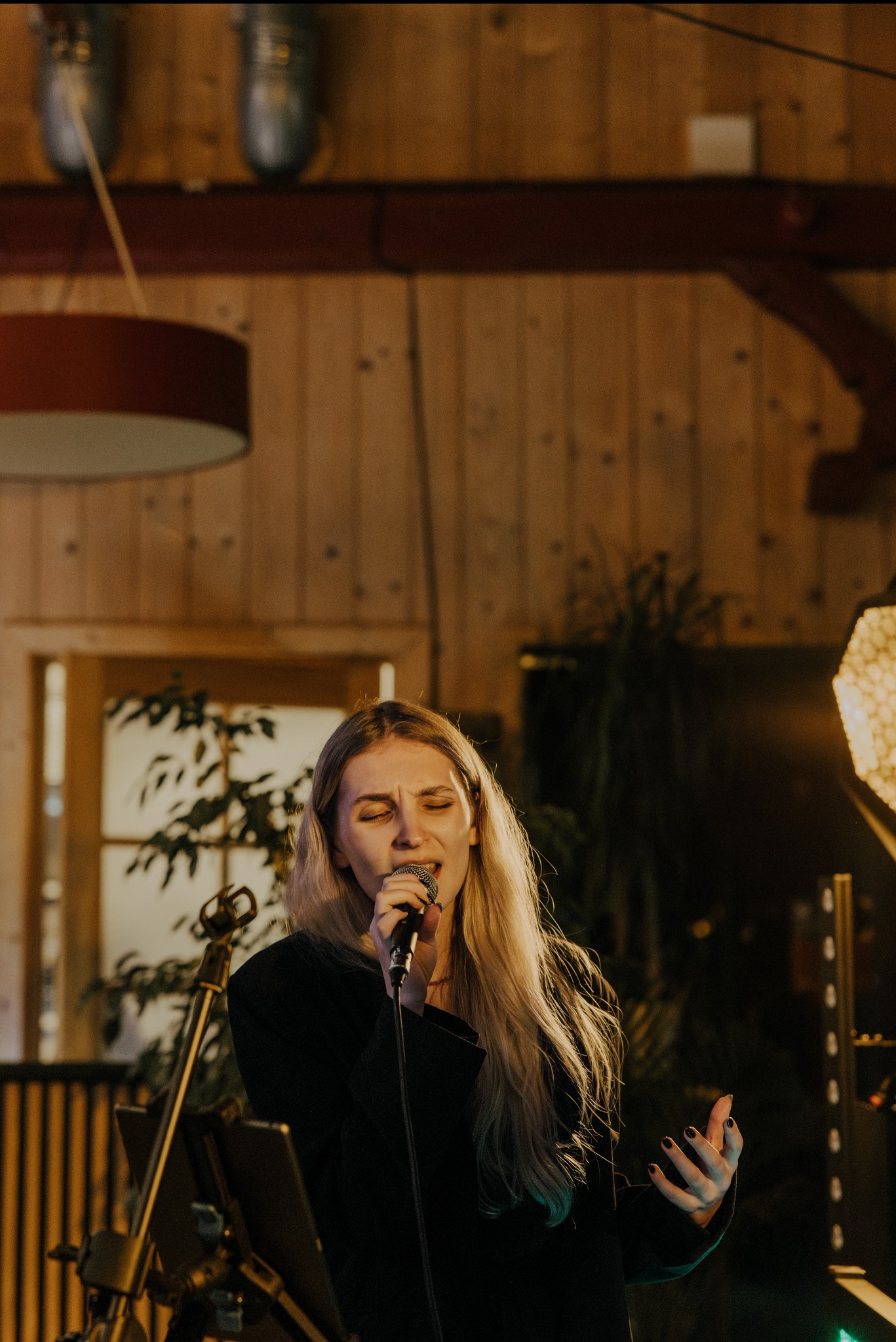 Blondwłosa wokalistka śpiewa do mikrofonu na scenie z drewnianym tłem, oświetlona ciepłym światłem reflektorów. Widoczny statyw mikrofonowy i fragment oświetlenia scenicznego.