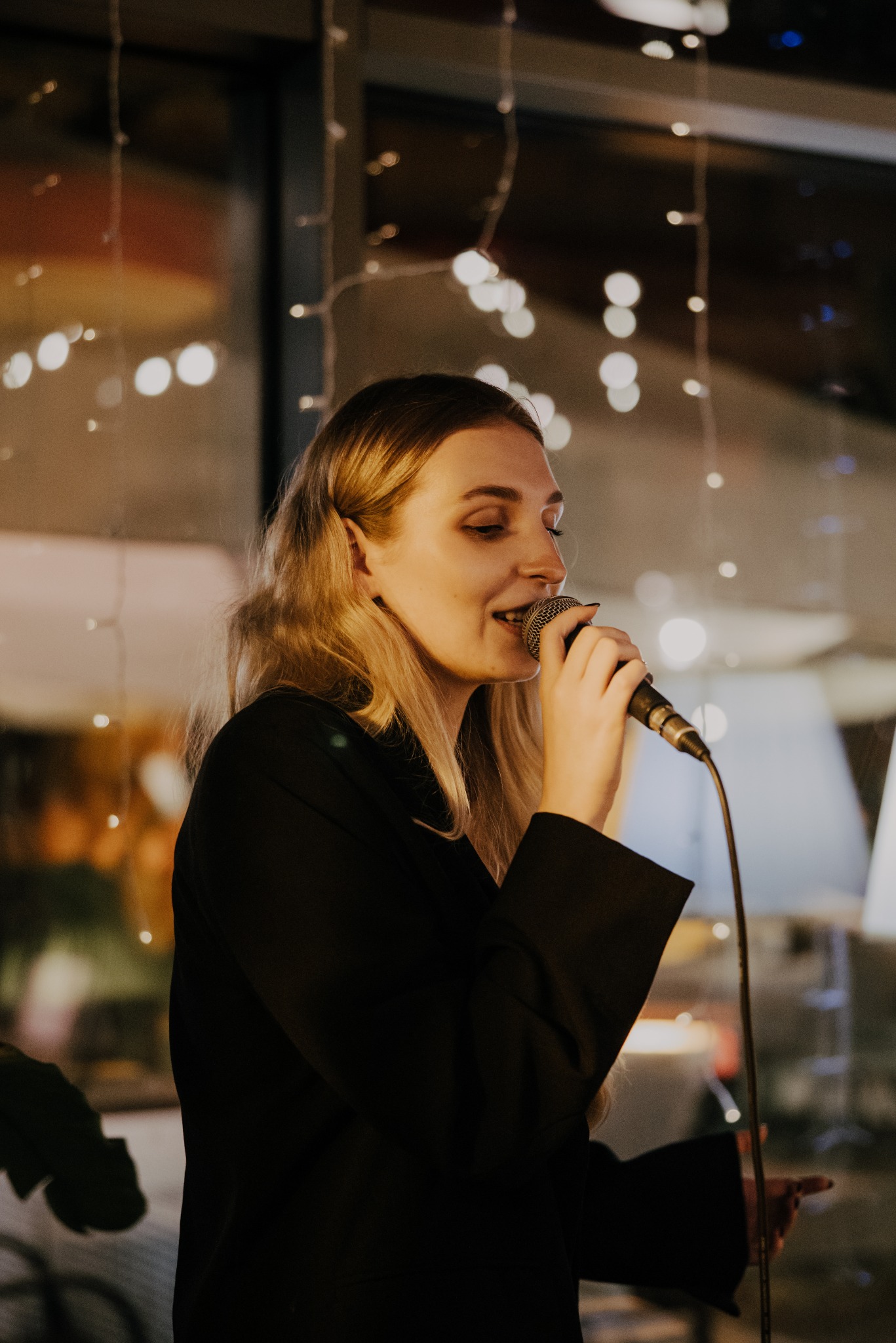 Blondwłosa wokalistka w czarnej marynarce śpiewa do mikrofonu na tle rozmytych światełek, tworząc intymną atmosferę koncertu.