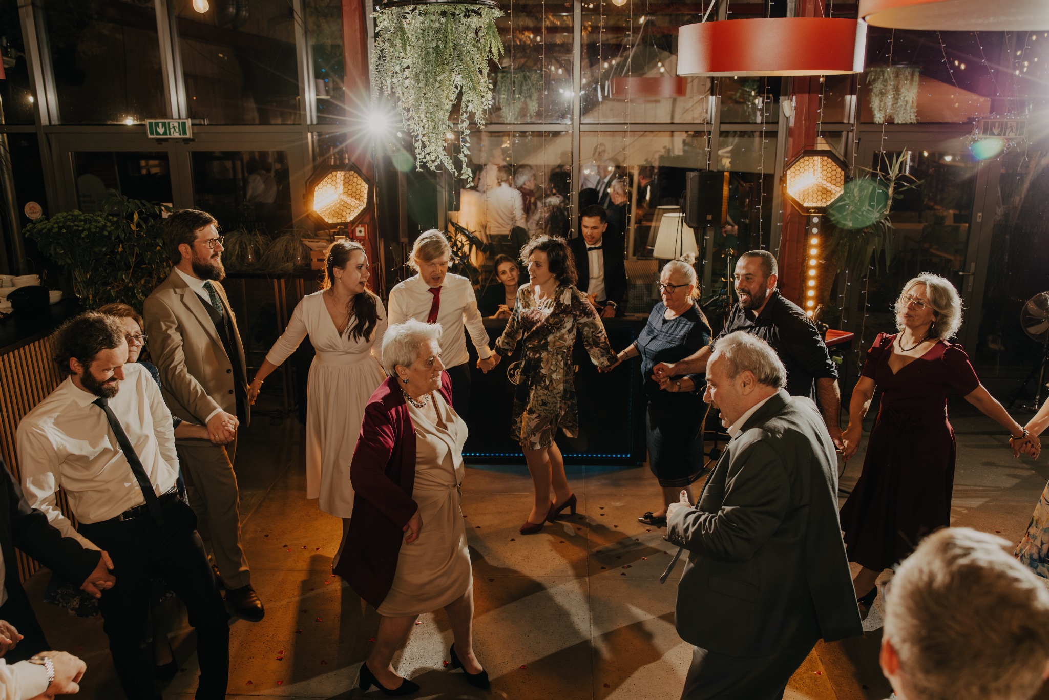 Radosny korowód gości weselnych trzymających się za ręce, w tle dekoracje świetlne i roślinne, parkiet z refleksami światła, ciepła atmosfera.