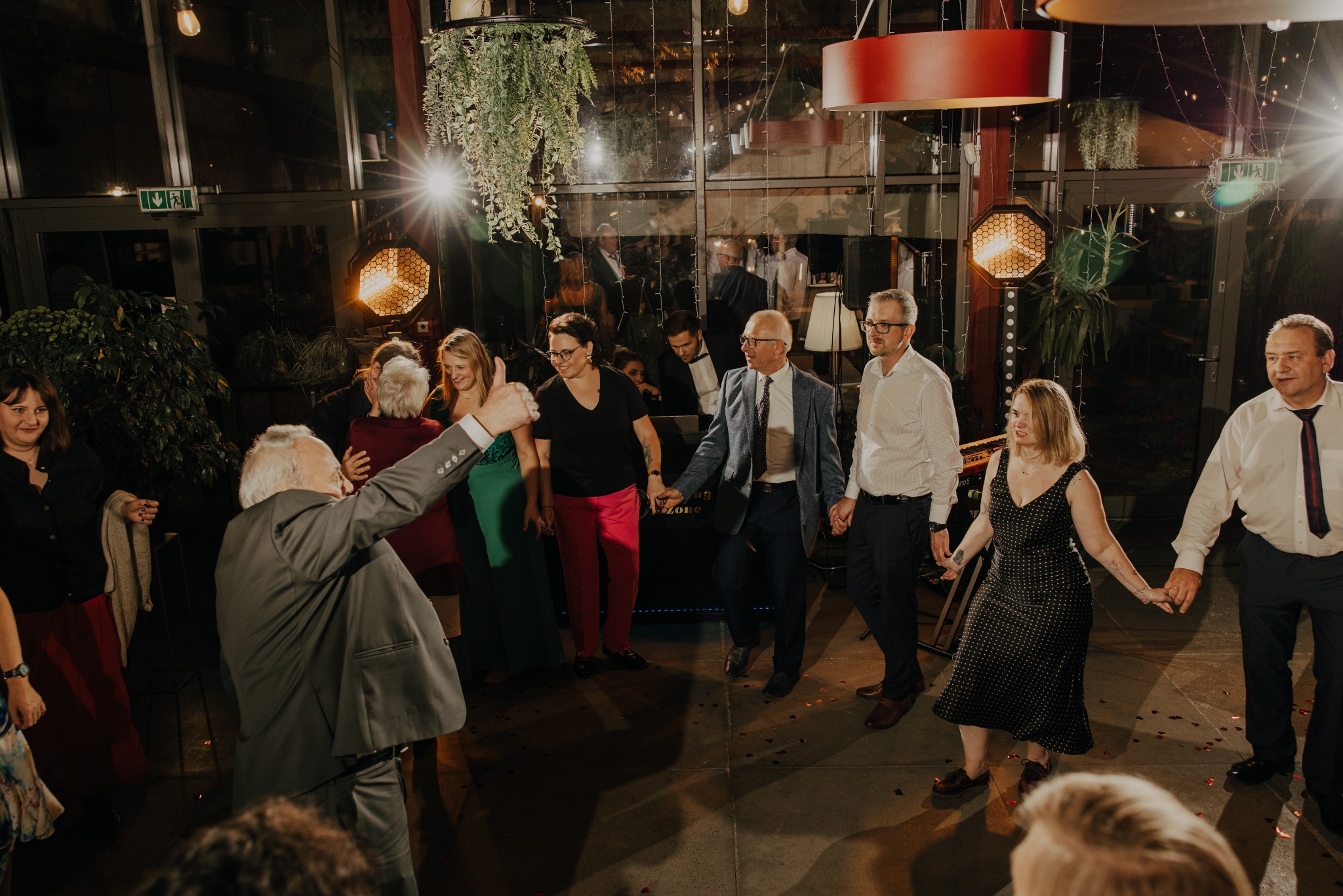 Rozświetlona sala balowa z tańczącymi gośćmi, oświetleniem eventowym i dekoracjami roślinnymi. W tle widoczny zespół muzyczny.