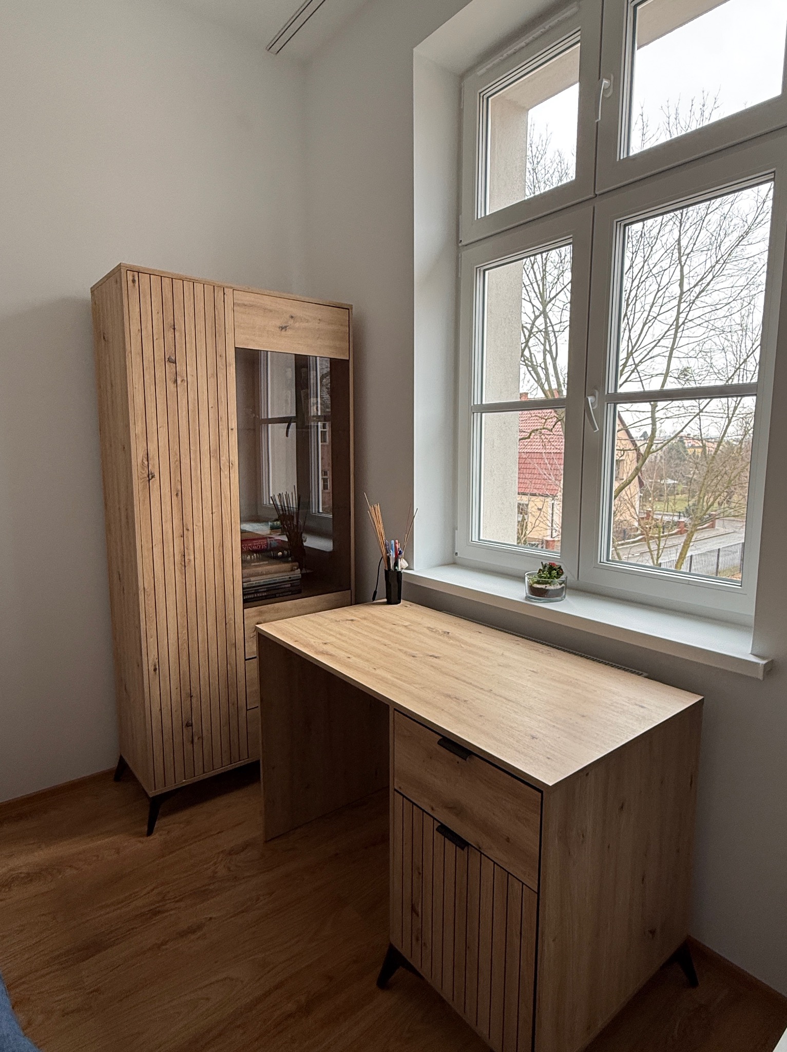 Jasne biuro z drewnianym biurkiem i regałem przy oknie. Minimalistyczny wystrój, naturalne światło. Na parapecie roślina w szklanym naczyniu.