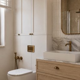 Projektant Wnętrz Studio RETRO Design - Patrycja Wawrzyniak - Elegancka łazienka z białymi szafkami, drewnianą szafką pod umywalkę, złotymi akcentami i marmurowym blatem. Widoczna podwieszana toaleta i okno z żaluzjami.