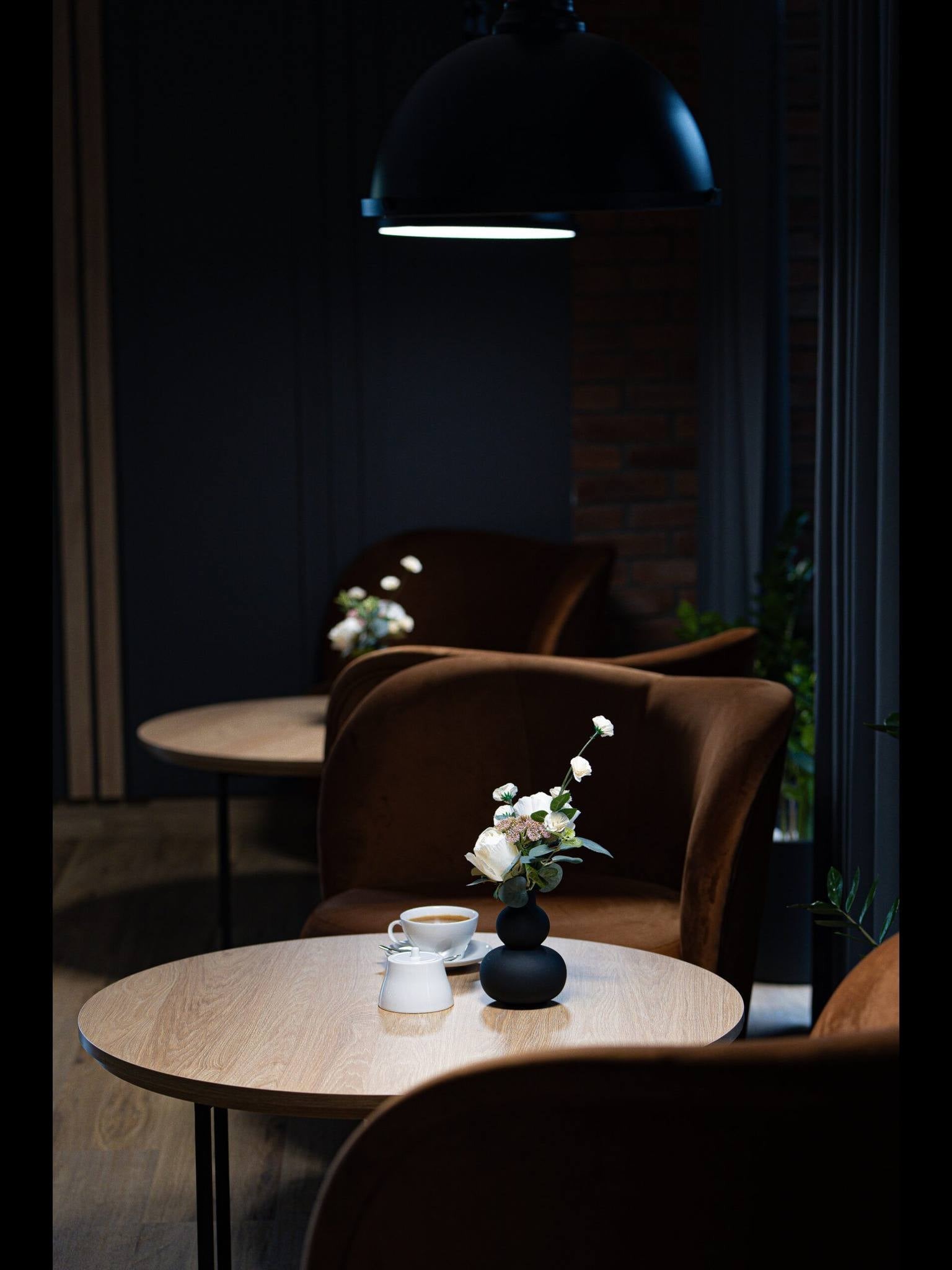 Kawiarniany stolik z bukietem i filiżanką kawy, brązowe fotele w tle, oświetlenie punktowe. Przytulne wnętrze kawiarni z ceglaną ścianą i ciemnymi elementami.