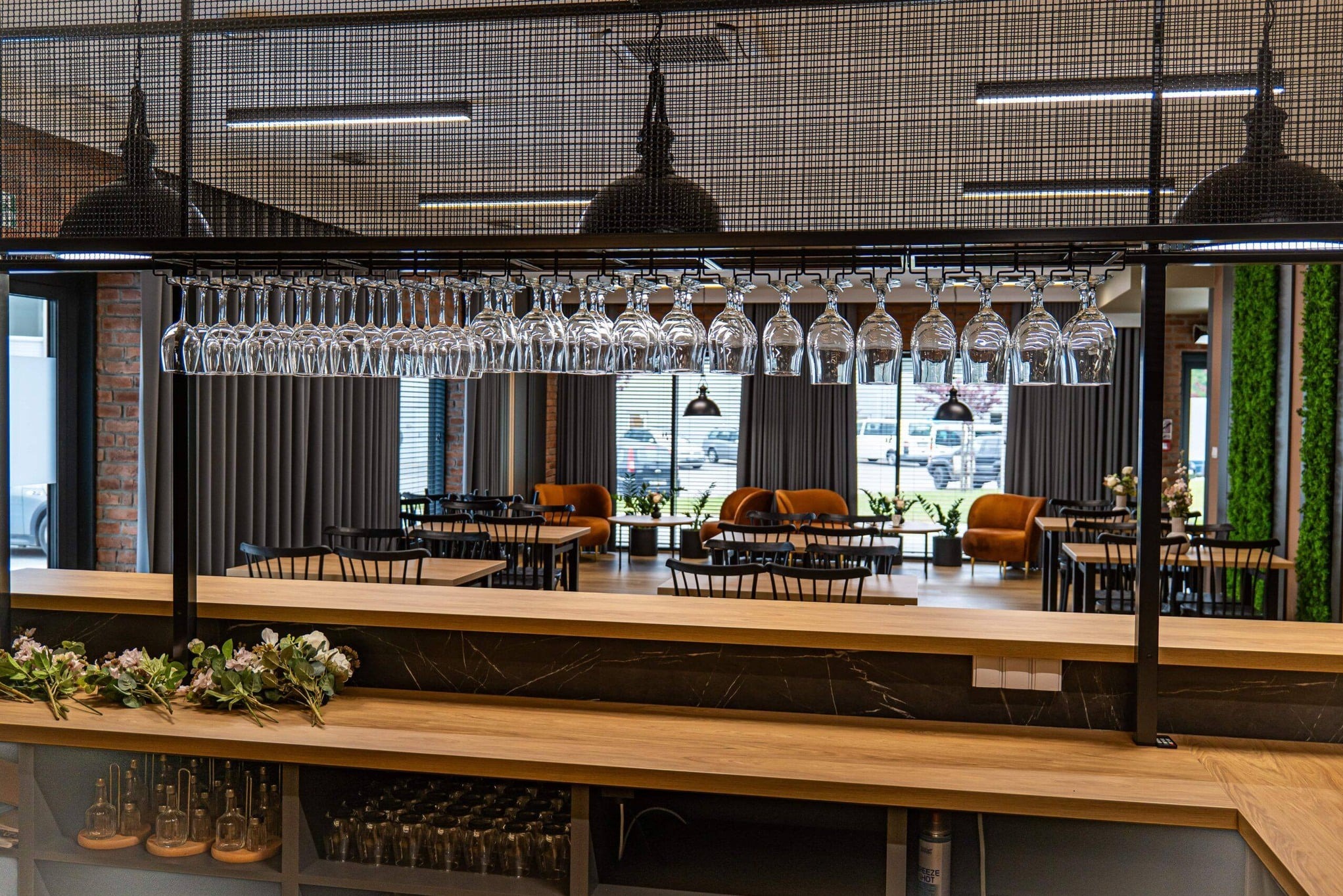 Nowoczesne wnętrze restauracji z drewnianym barem, wiszącymi kieliszkami i pomarańczowymi fotelami w tle. Minimalistyczny design z elementami industrialnymi.