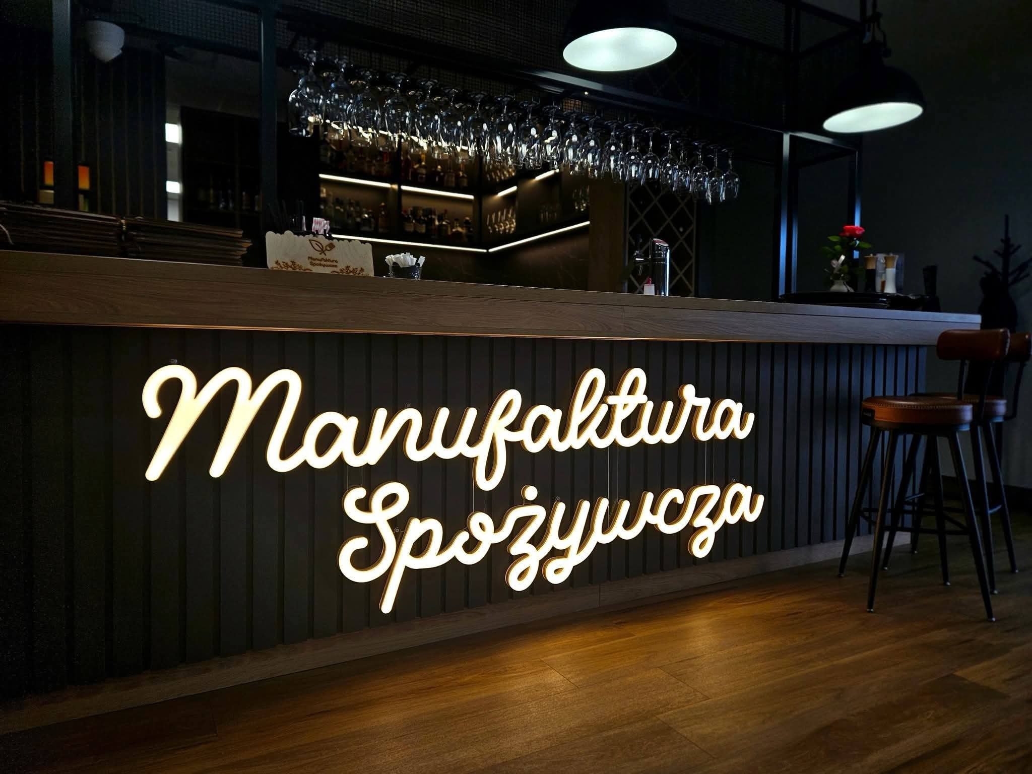 Podświetlany neon 'Manufaktura Spożywcza' na barze z ciemnego drewna, szklanki wiszące nad barem, krzesła barowe w tle, ciepłe oświetlenie, nowoczesny wystrój.