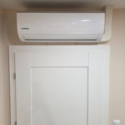 KW KlimaVent Wojciech Świat - Biały klimatyzator Kaisai zamontowany nad białymi drzwiami w jasnym wnętrzu. Widoczna instalacja odprowadzająca. Minimalistyczny design.