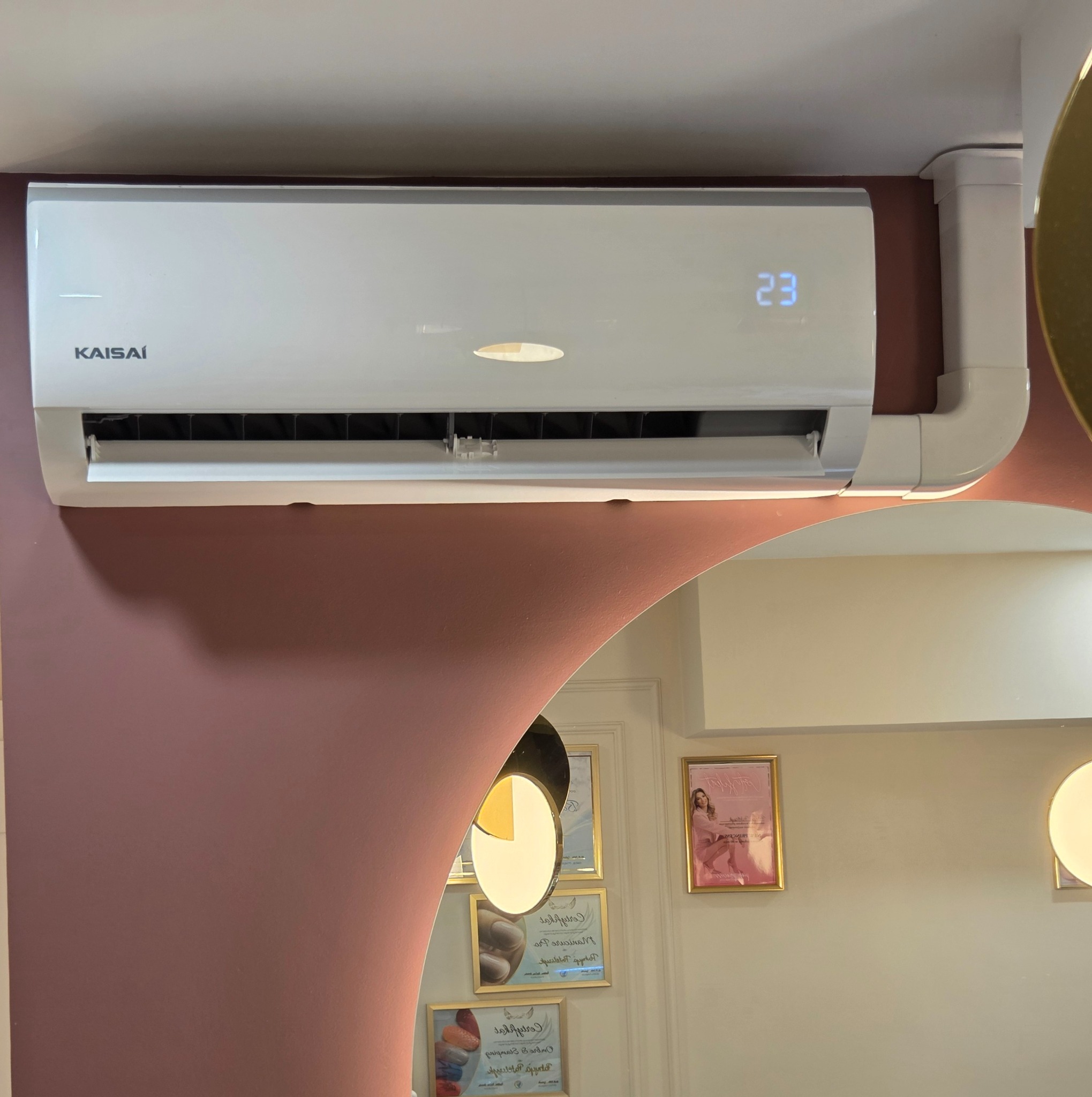 Biały klimatyzator Kaisai z wyświetlaczem temperatury 23°C, zamontowany na różowej ścianie w pomieszczeniu z lustrem i certyfikatami na ścianie.