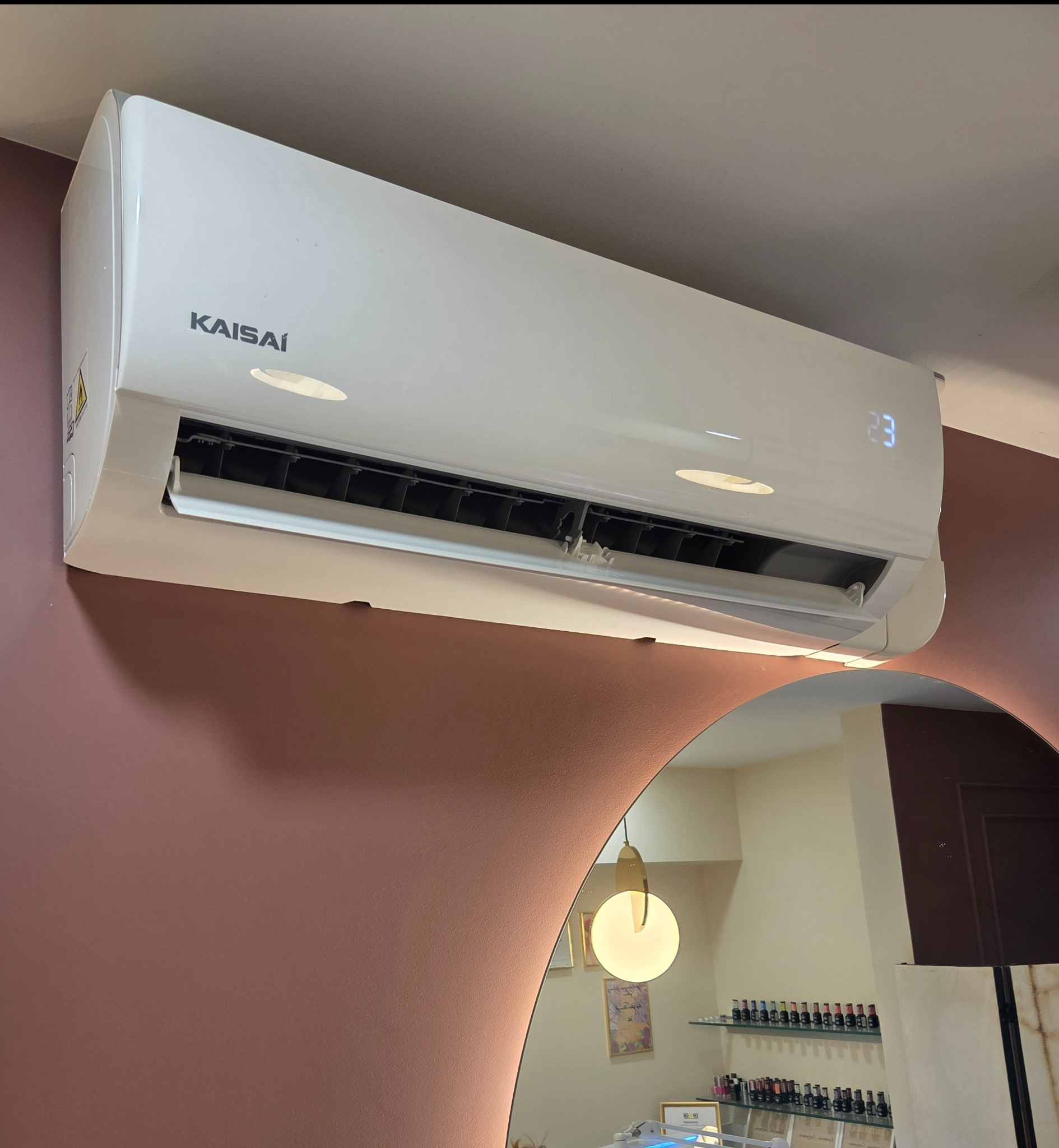 Biały klimatyzator Kaisai na ścianie w salonie kosmetycznym, widoczny w odbiciu lustra. Ustawiona temperatura 23 stopnie Celsjusza.