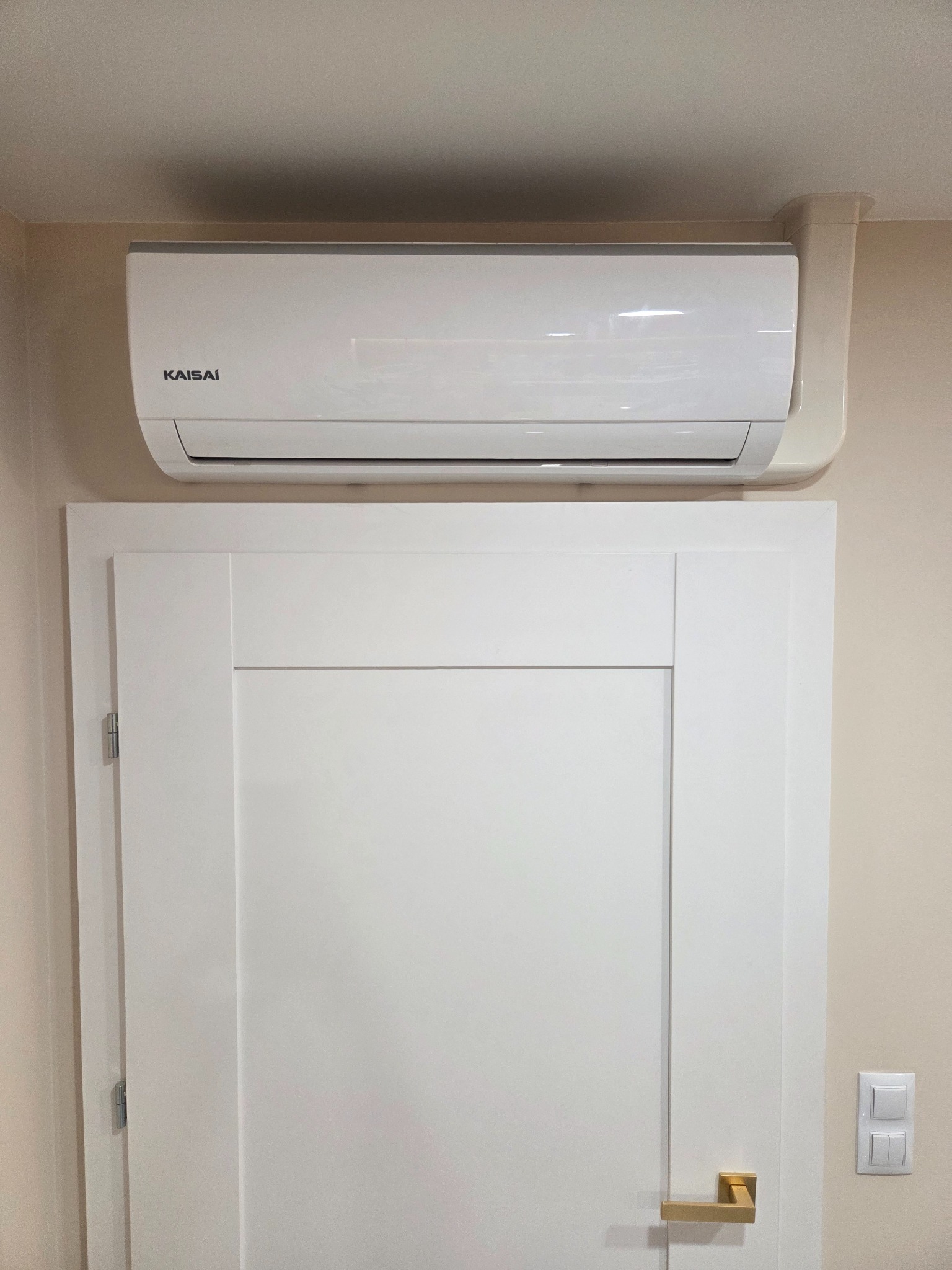 Biały klimatyzator Kaisai zamontowany nad białymi drzwiami w jasnym wnętrzu. Widoczna instalacja odprowadzająca. Minimalistyczny design.